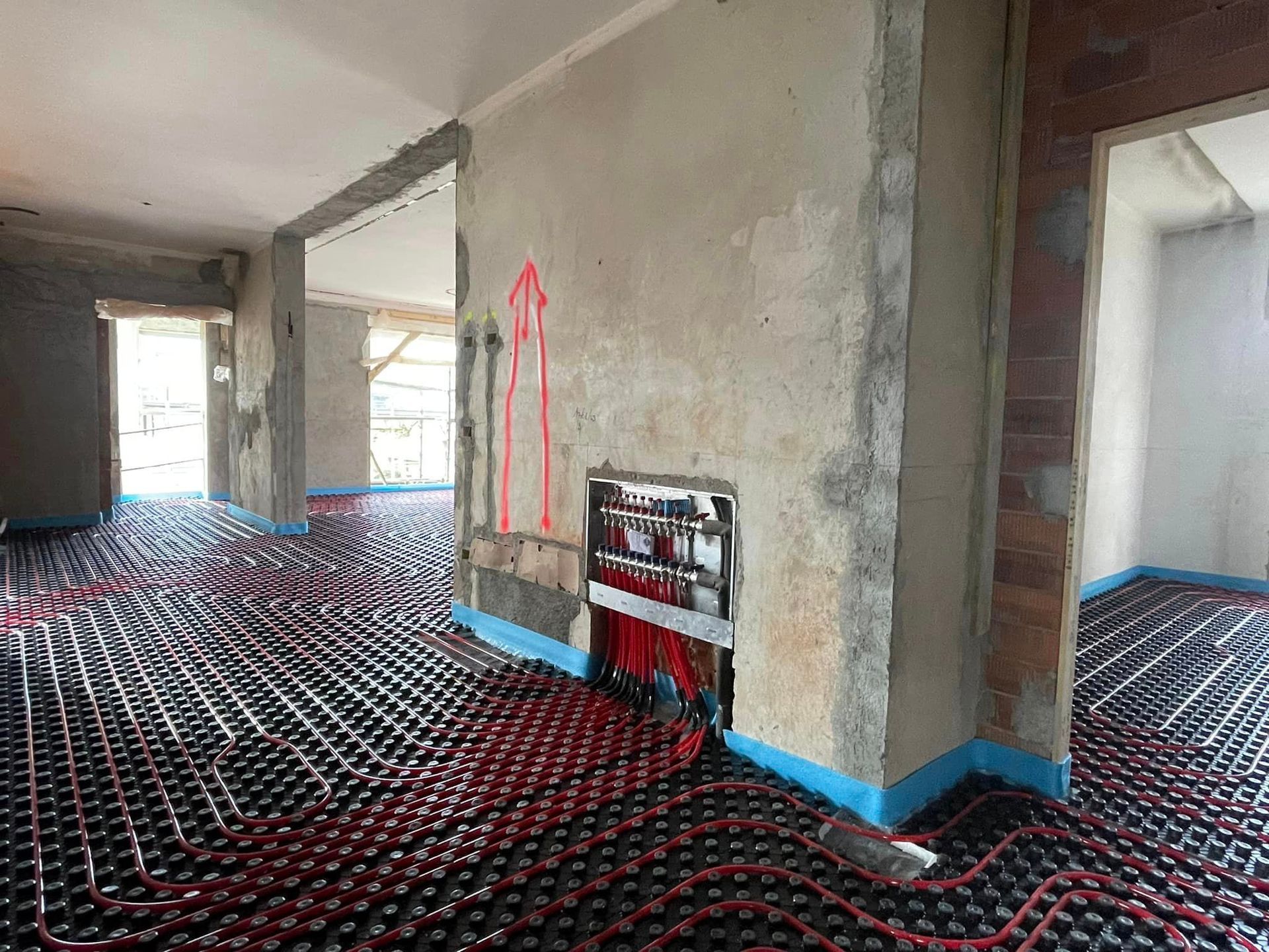 Stanza non rifinita con pareti e tubi del riscaldamento a pavimento a vista, nastro adesivo blu alla base e un segno rosso su una parete.