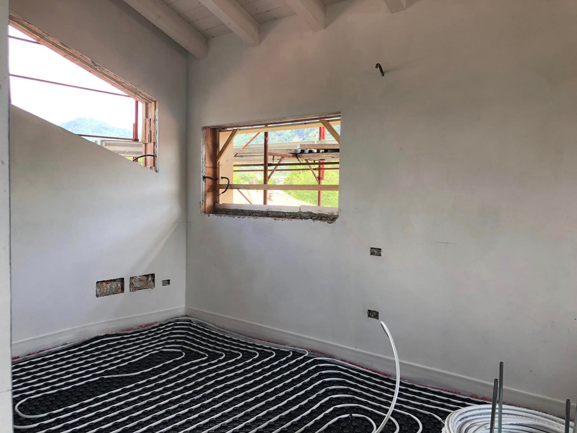 Stanza vuota e non rifinita con pareti bianche, finestre a vista e tubi del riscaldamento a pavimento coperti da tubi bianchi e neri.