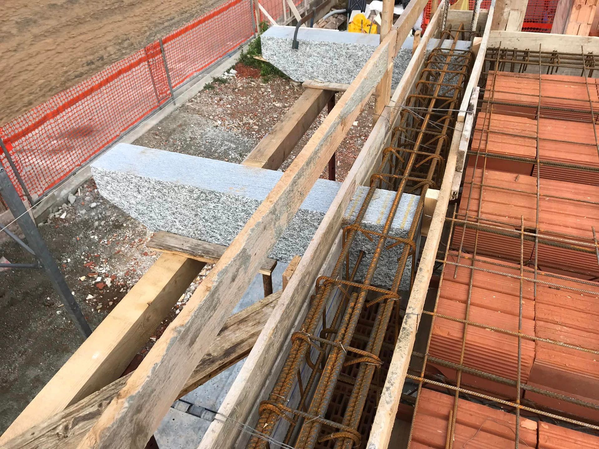 Cantiere edile con travi di legno, barre d'armatura e casseforme per il cemento, accanto a una recinzione di sicurezza arancione.