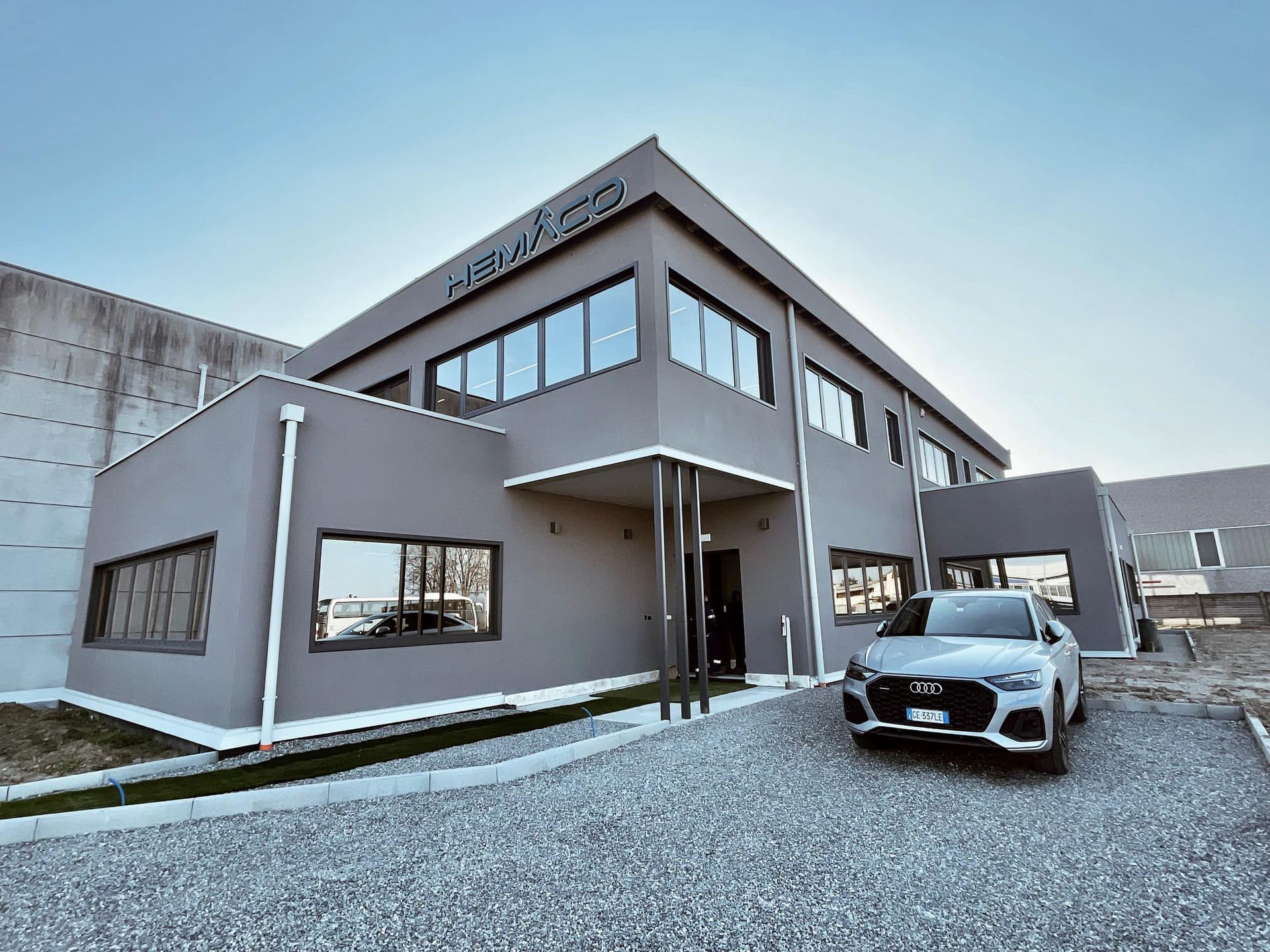 Moderno edificio grigio a due piani con un'auto parcheggiata su un piazzale di ghiaia sotto un cielo limpido