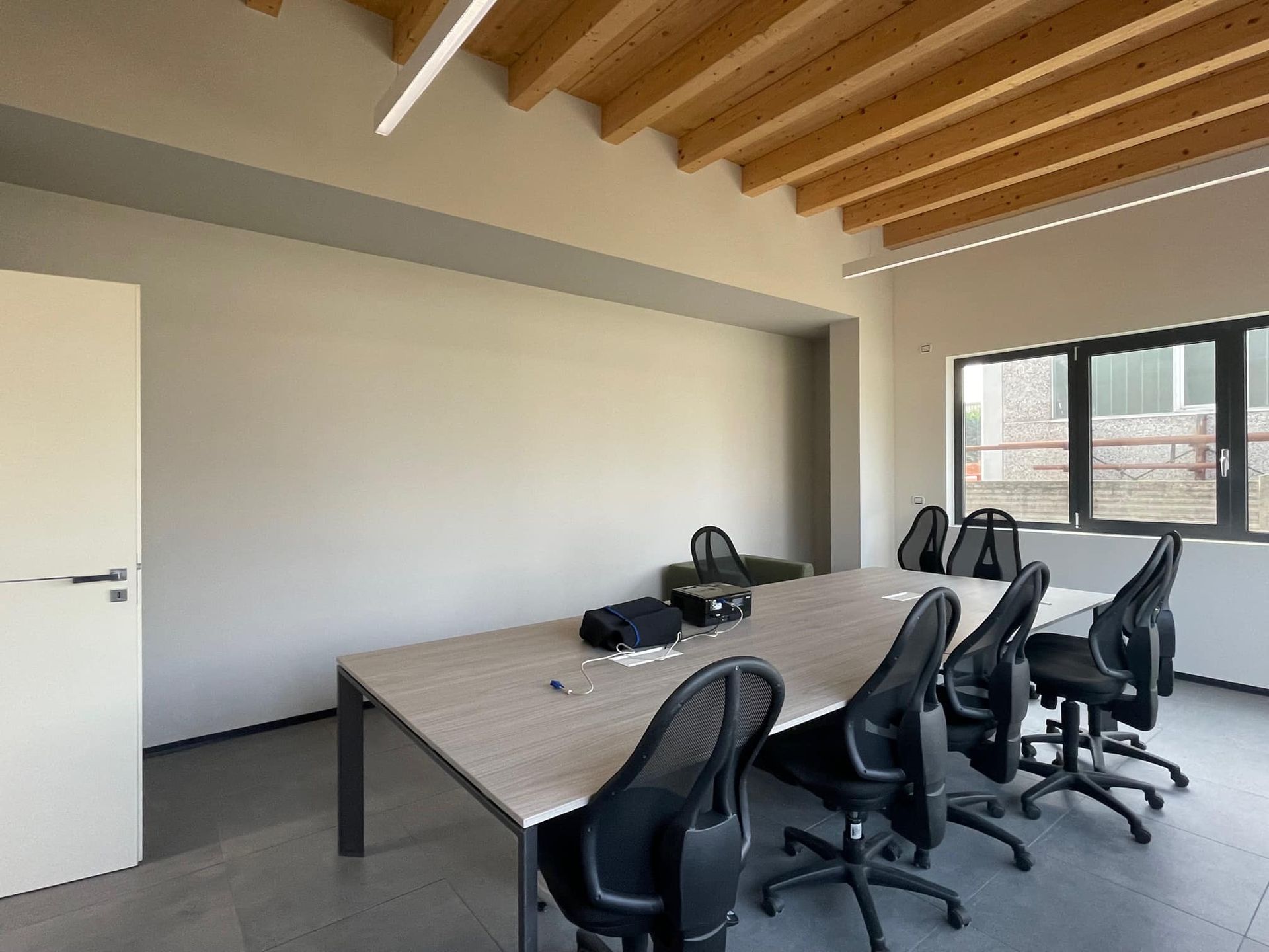 Sala riunioni moderna con sedie disposte attorno a un lungo tavolo, pareti bianche, soffitto in legno e una finestra.