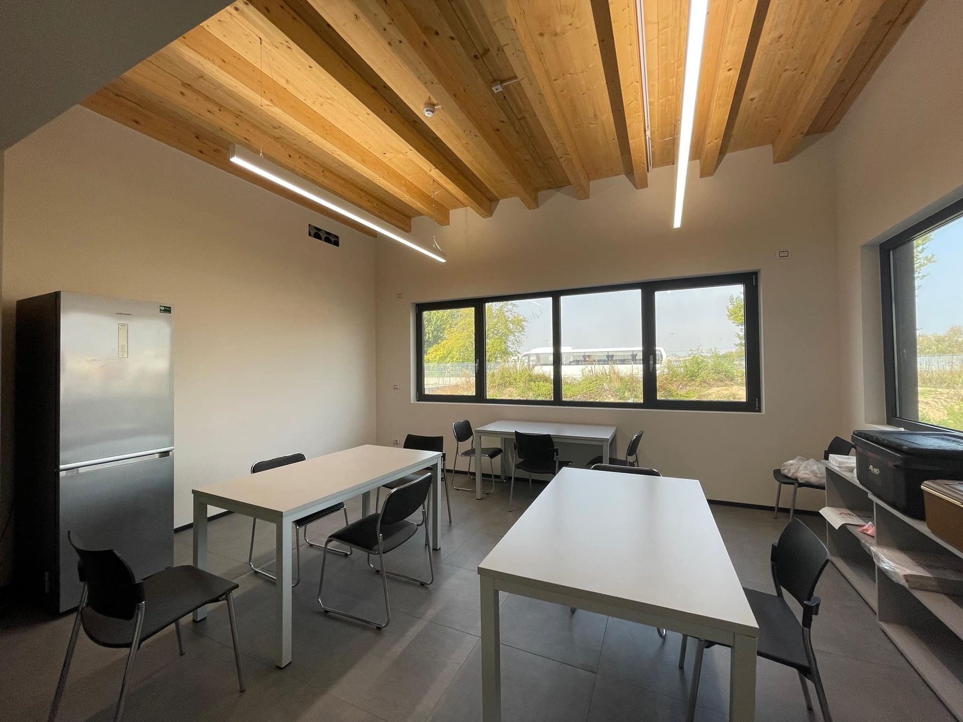 Camera luminosa con soffitto in legno, ampie finestre, tavoli, sedie e frigorifero.