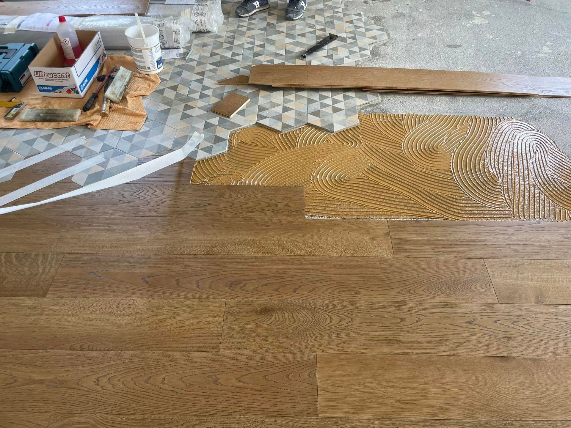 Pavimento in legno massello parzialmente installato con zona adesiva non rifinita e attrezzi in una stanza in fase di ristrutturazione.