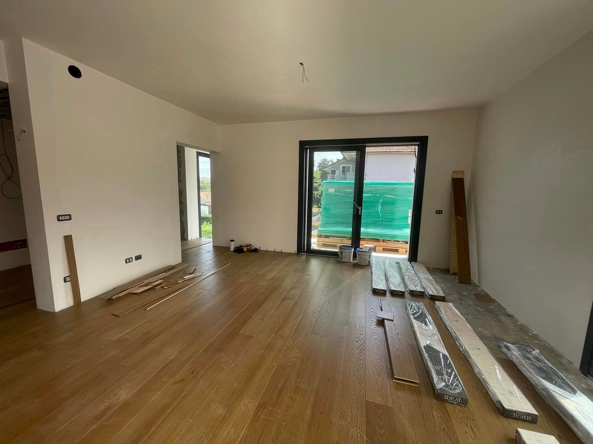 Stanza vuota in fase di ristrutturazione con pavimento in legno, pareti bianche e porte scorrevoli in vetro che danno su un balcone.