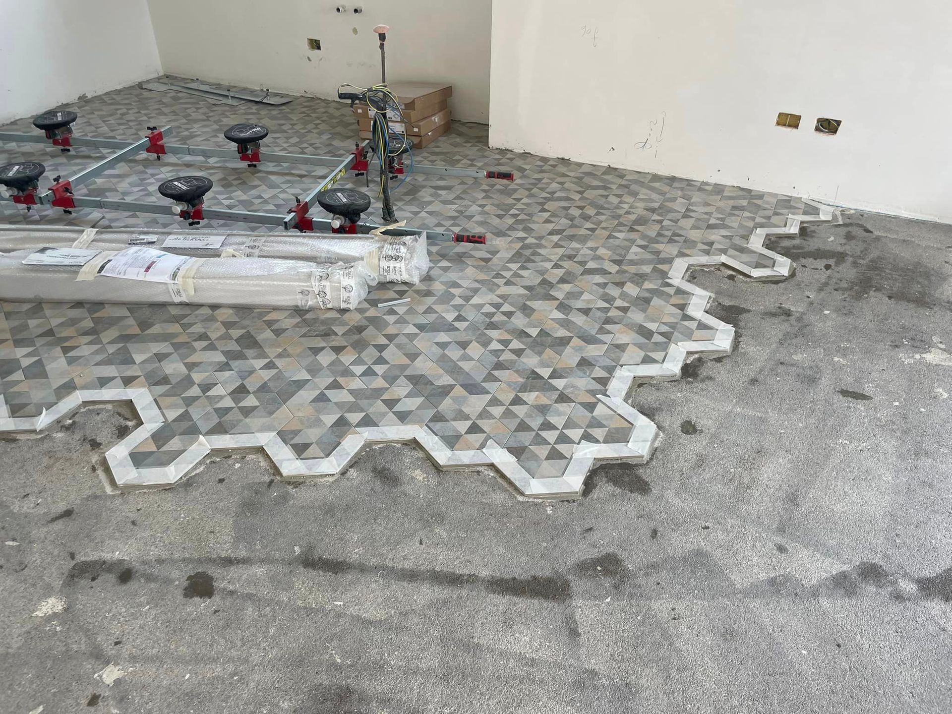 Cantiere edile con pavimentazione grigia a motivi geometrici in fase di posa accanto a un muro, con attrezzi e distanziatori a terra.