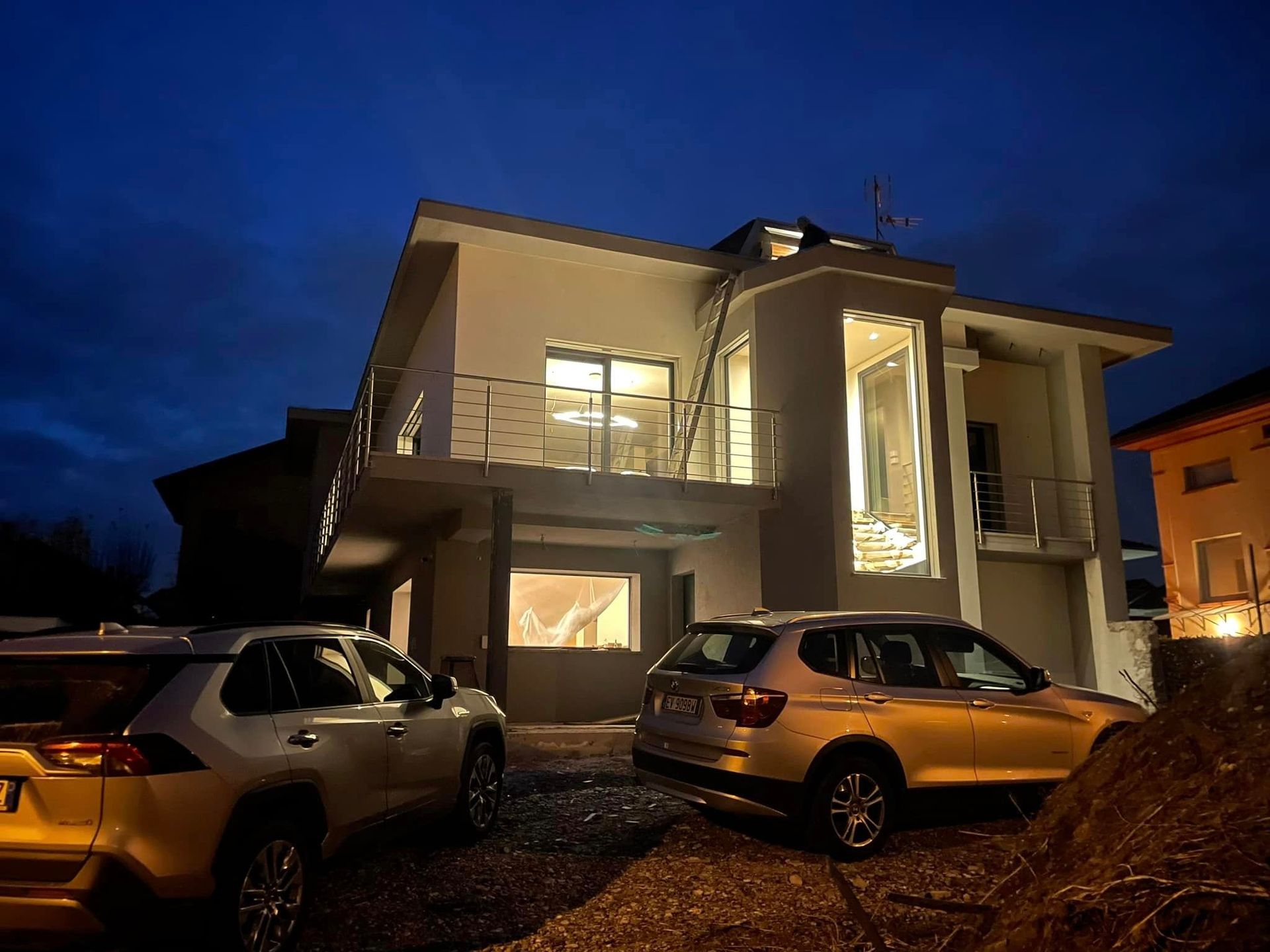 Casa moderna a due piani al crepuscolo, con luci interne calde e due SUV parcheggiati nel vialetto.