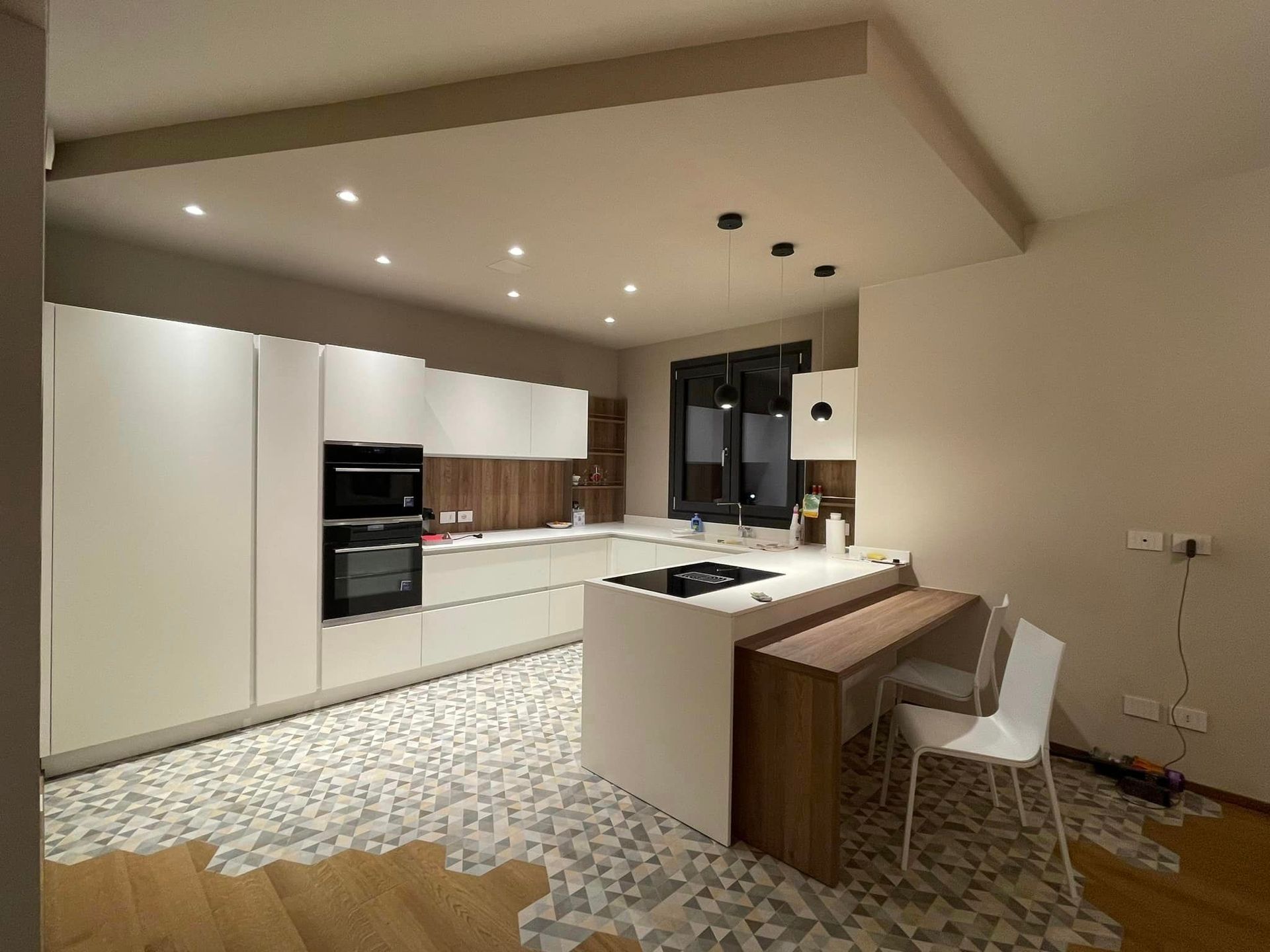 Cucina moderna bianca con dettagli in legno, pavimento piastrellato con motivi decorativi, isola e sgabelli da bar sotto faretti a incasso.