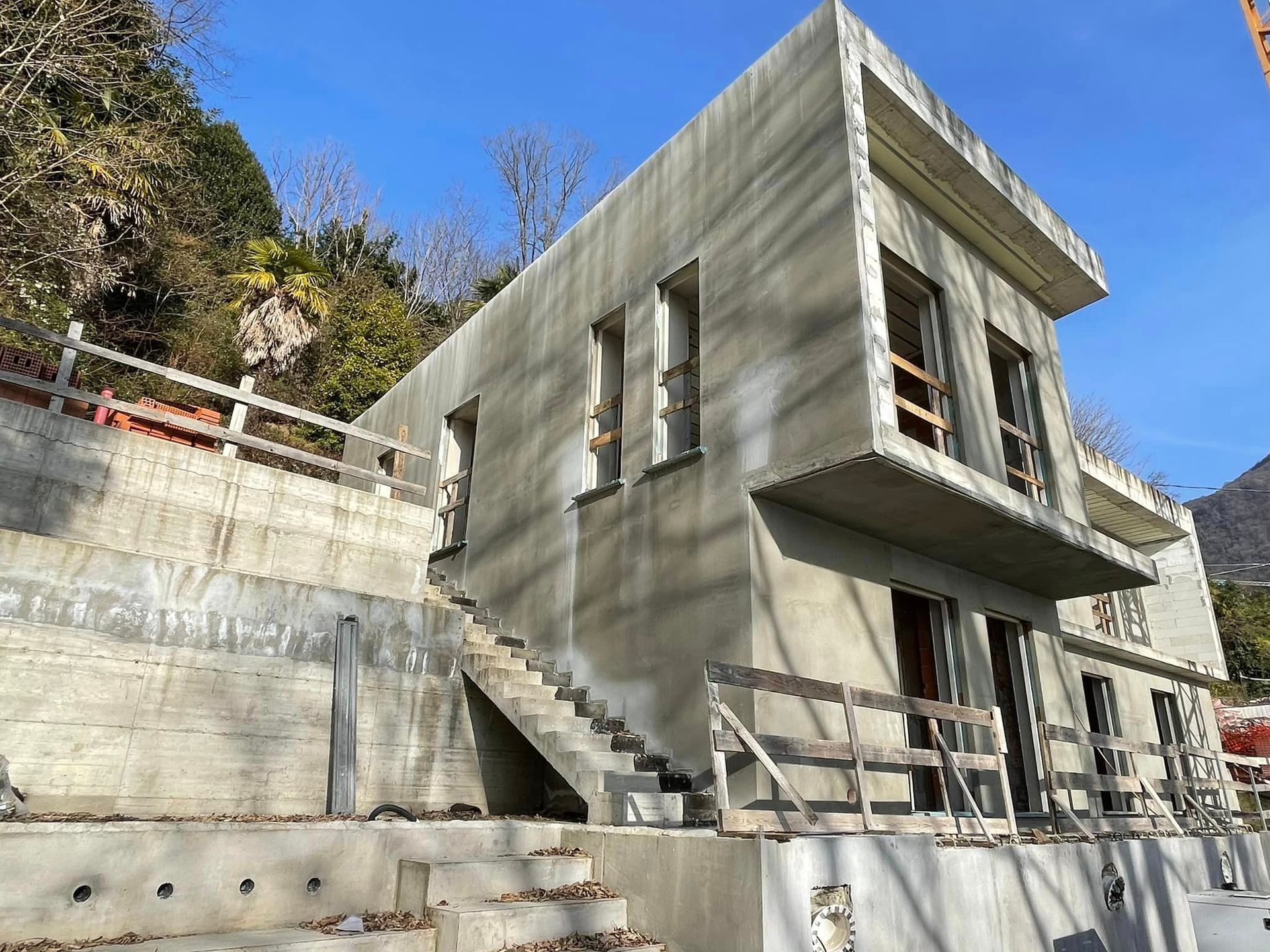 Casa in cemento in costruzione su una collina, con scala esterna e cielo azzurro sopra.