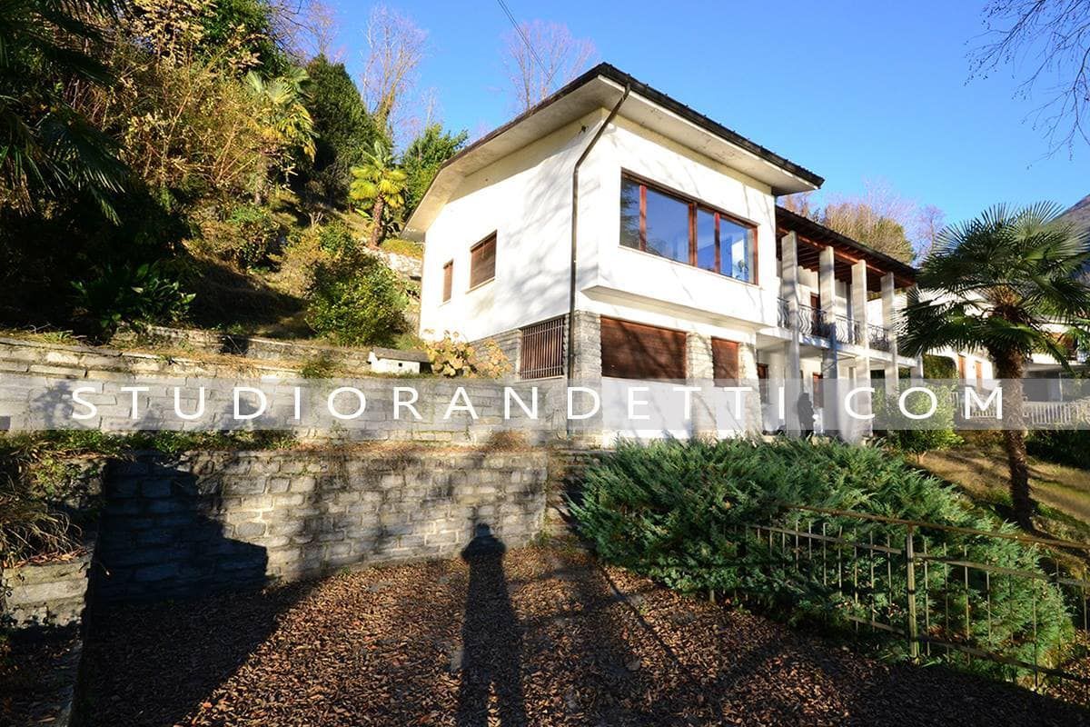 Casa moderna bianca su una collina con muro in pietra, grandi finestre e vegetazione circostante sotto un cielo azzurro.