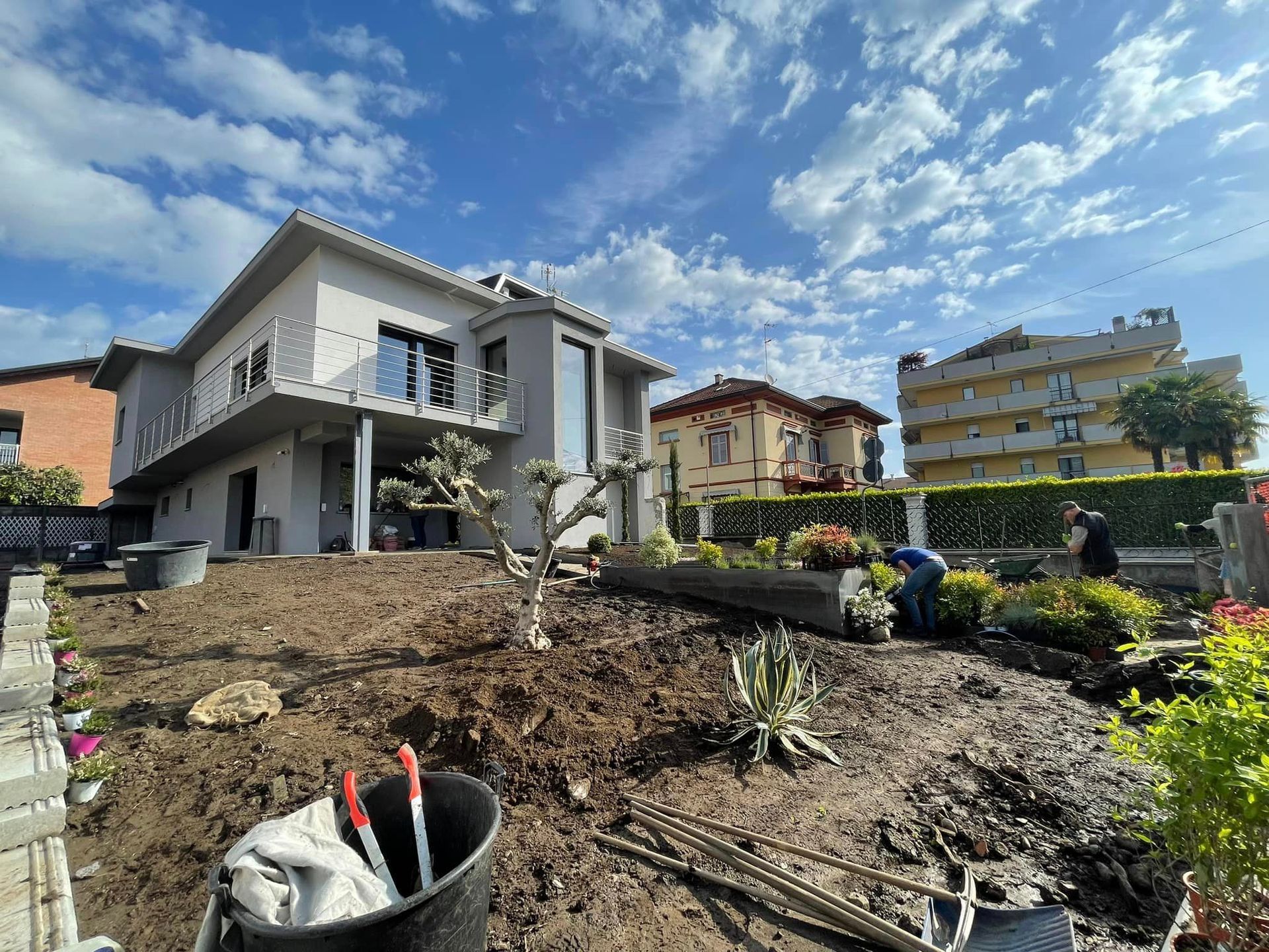Giardino con terra appena smossa di fronte a una casa grigia sotto un cielo azzurro, con case vicine nelle vicinanze.