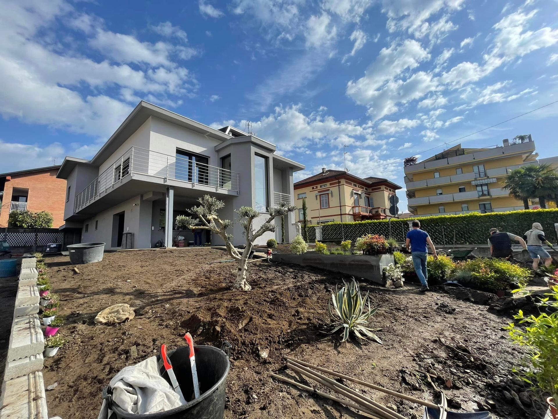 Scavo nel giardino antistante una casa moderna, con un operaio, cumuli di terra e lavori di giardinaggio sotto un cielo azzurro.