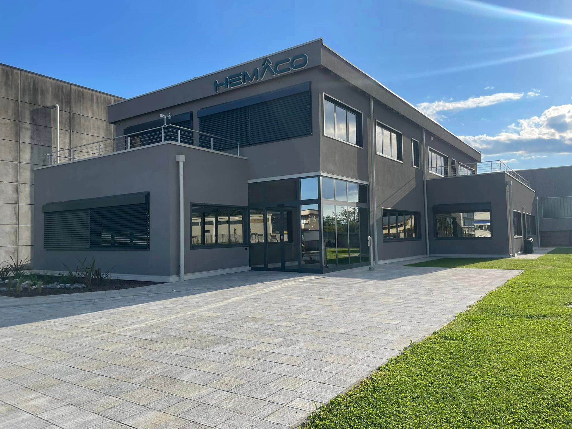Moderno edificio per uffici di colore grigio con ampie finestre, vialetto in pietra e prato verde sotto un cielo azzurro e soleggiato.