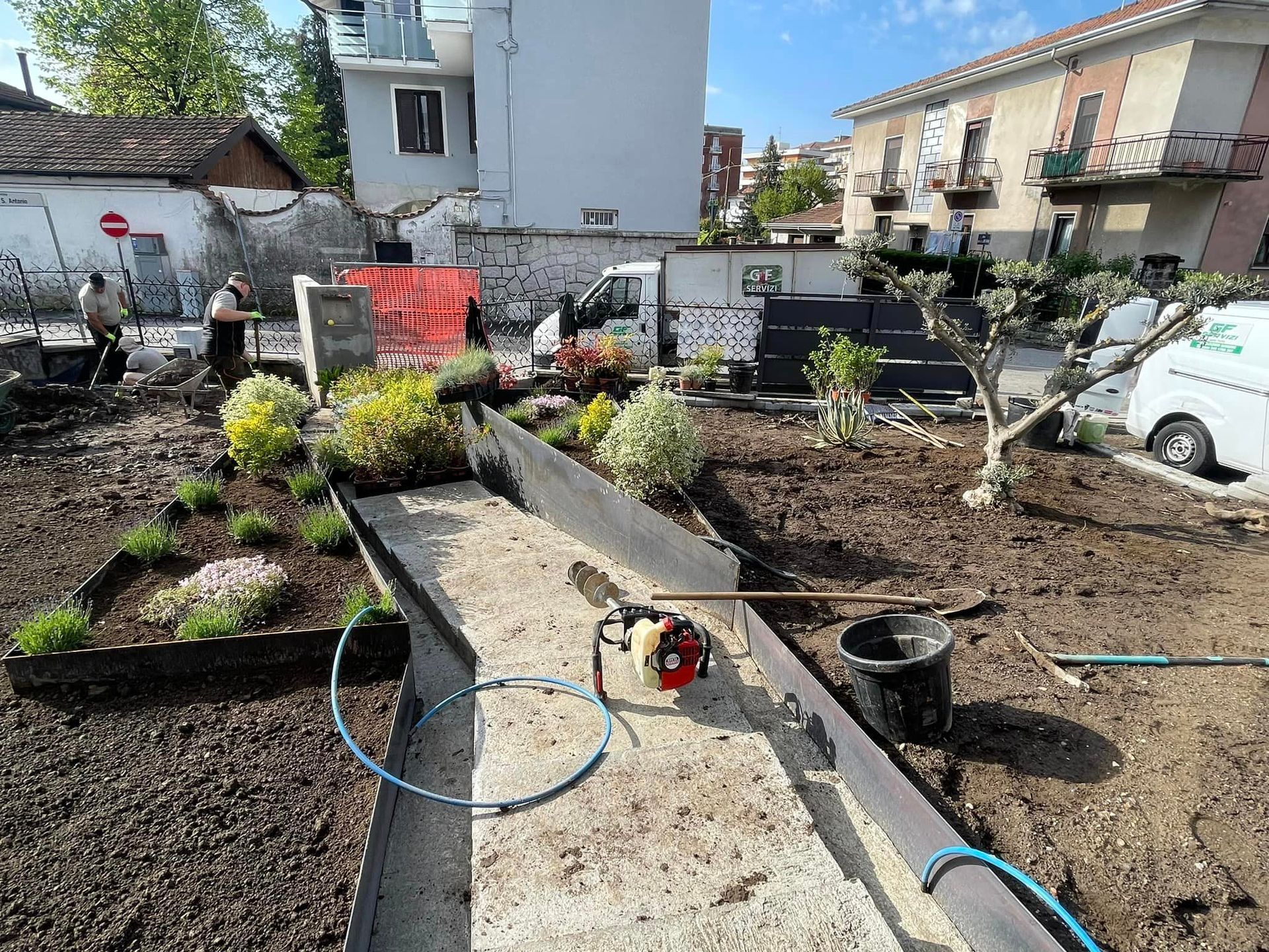 Giardino sul retro in fase di costruzione con vialetto in cemento, aiuole, attrezzi e auto parcheggiate nelle vicinanze.