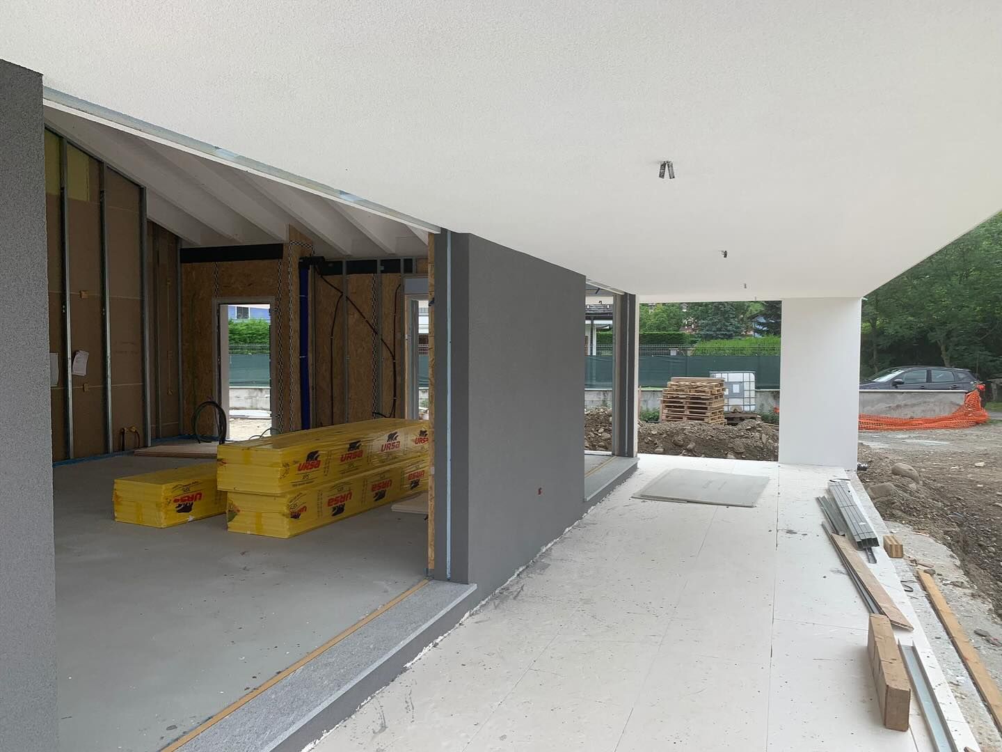 Casa moderna parzialmente costruita con struttura a vista, materiali sovrapposti e vialetto o patio coperto.