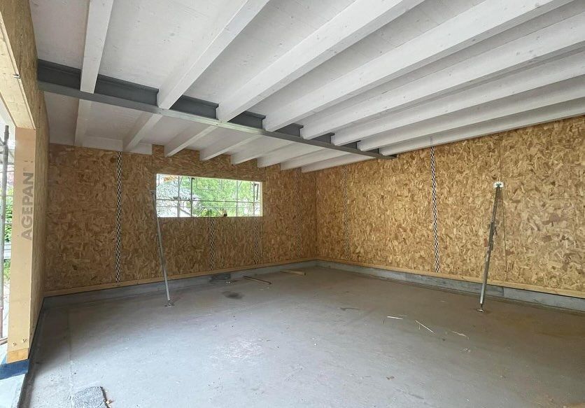 Stanza non rifinita con pareti in compensato, travi a vista sul soffitto, pavimento in cemento e una piccola finestra.