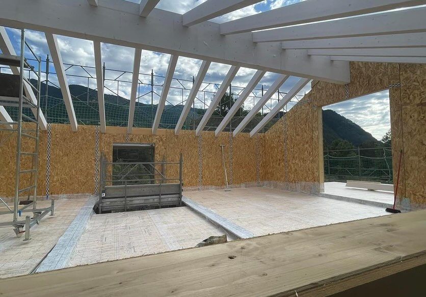 Interno di una casa parzialmente costruita con travi a vista, pareti non rifinite e vista sulle montagne attraverso ampie aperture.
