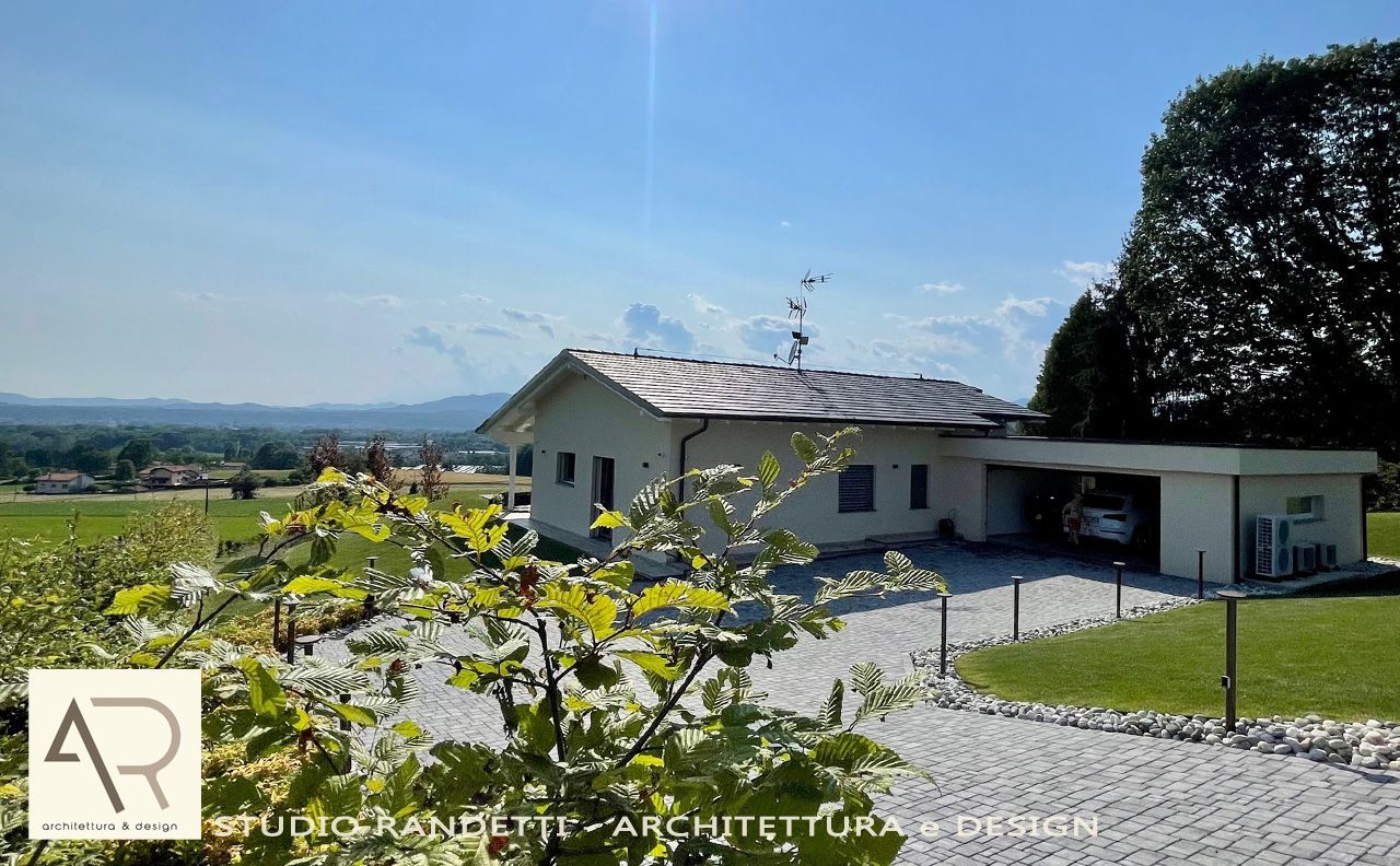 Casa moderna bianca con tetto grigio, vialetto di ghiaia e collina verdeggiante sotto un cielo azzurro limpido.