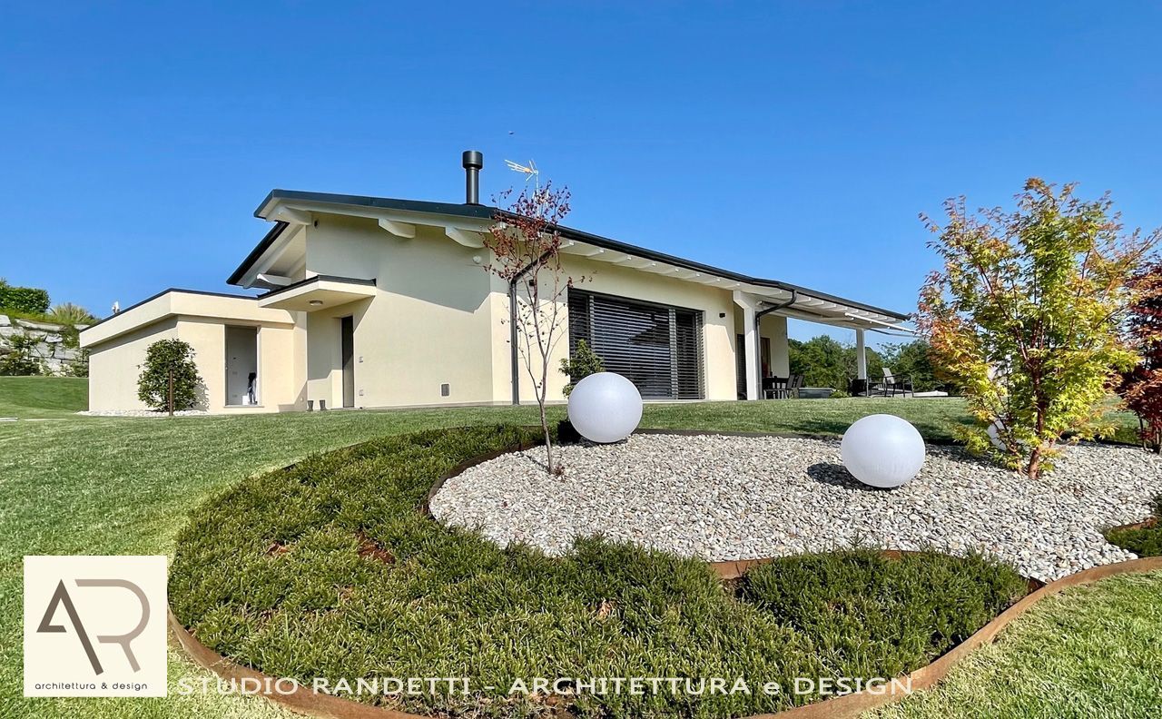 Casa moderna color beige con un giardino anteriore curato, aiuola di ghiaia e sculture sferiche sotto un cielo azzurro.