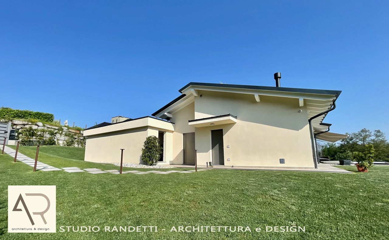 Casa moderna color beige su una collina erbosa sotto un cielo azzurro, con un piccolo patio e tetto spiovente.