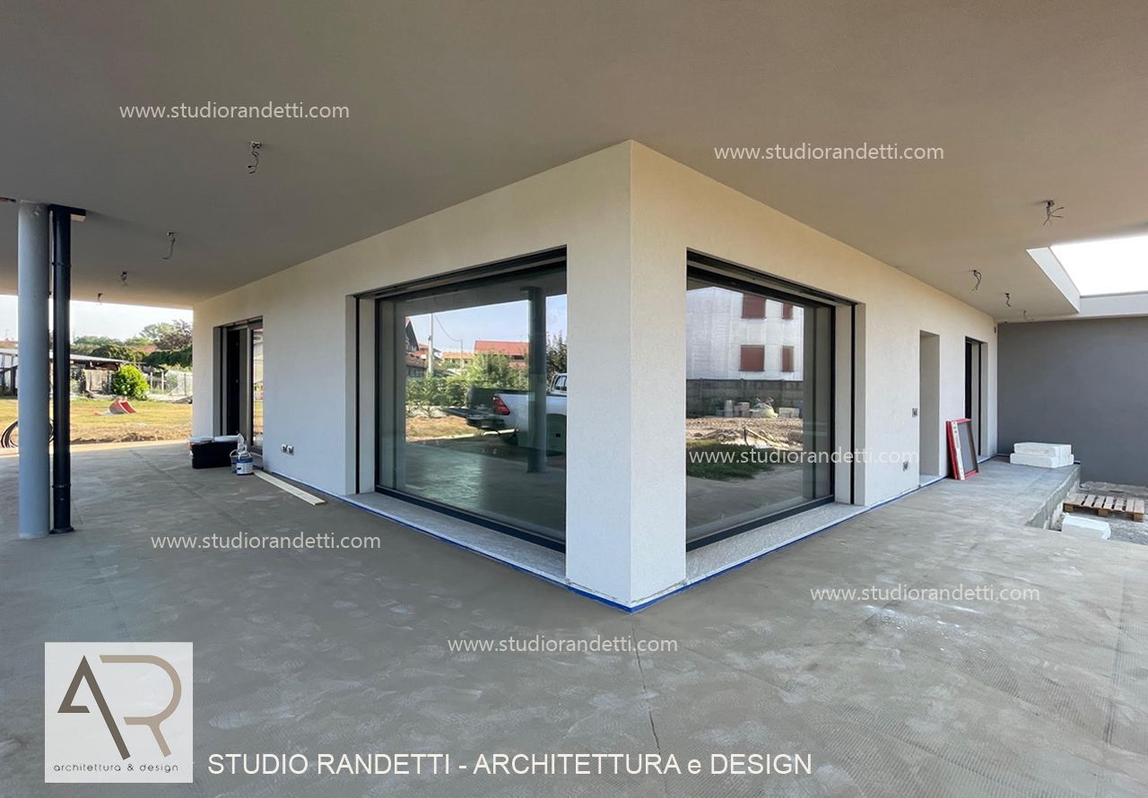 Esterno di una casa moderna con ampie finestre a tutta altezza, patio coperto e terrazza in cemento.
