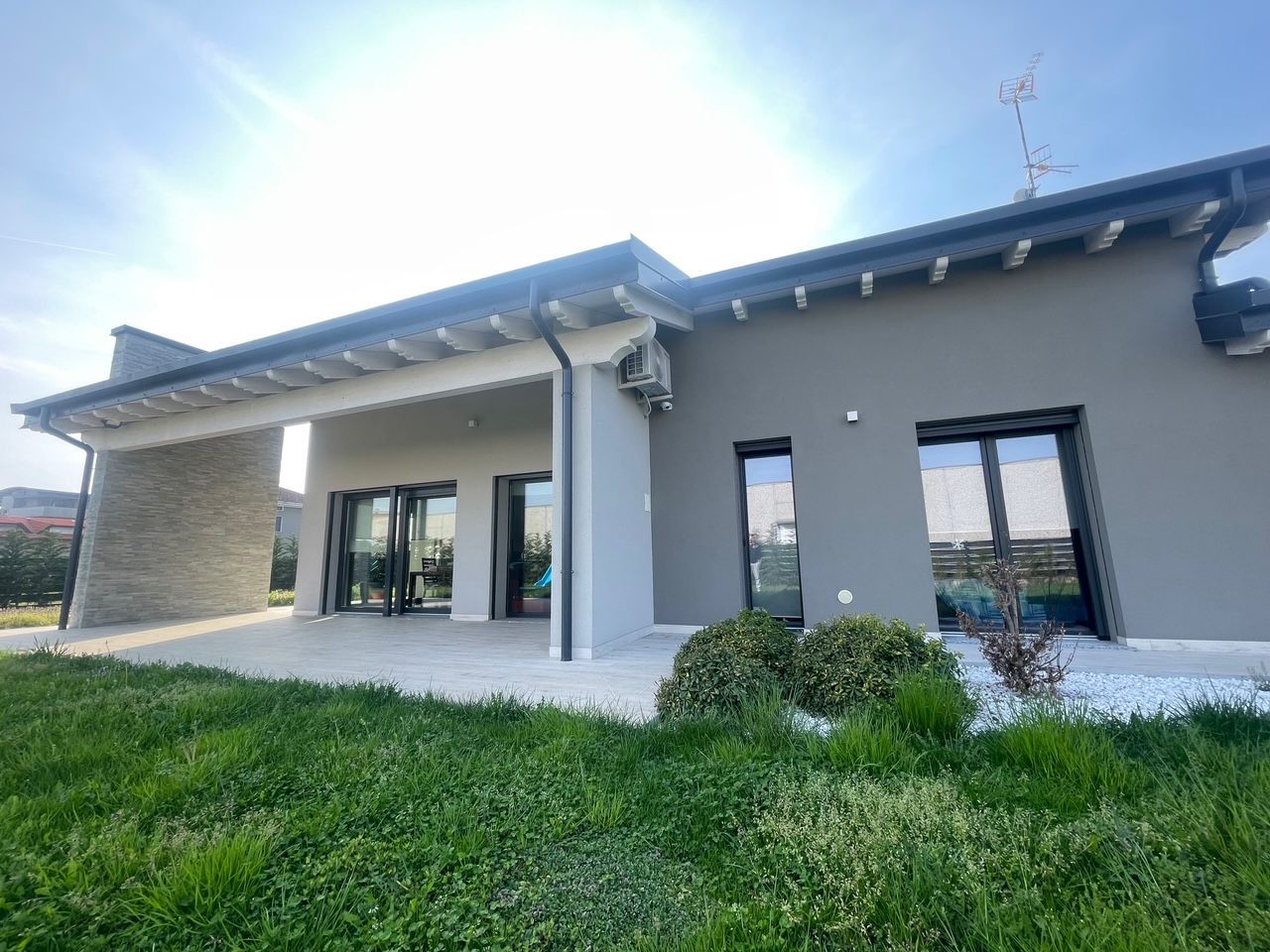 Esterno moderno di una casa grigia con ampie finestre, patio coperto e prato verde sotto un sole splendente