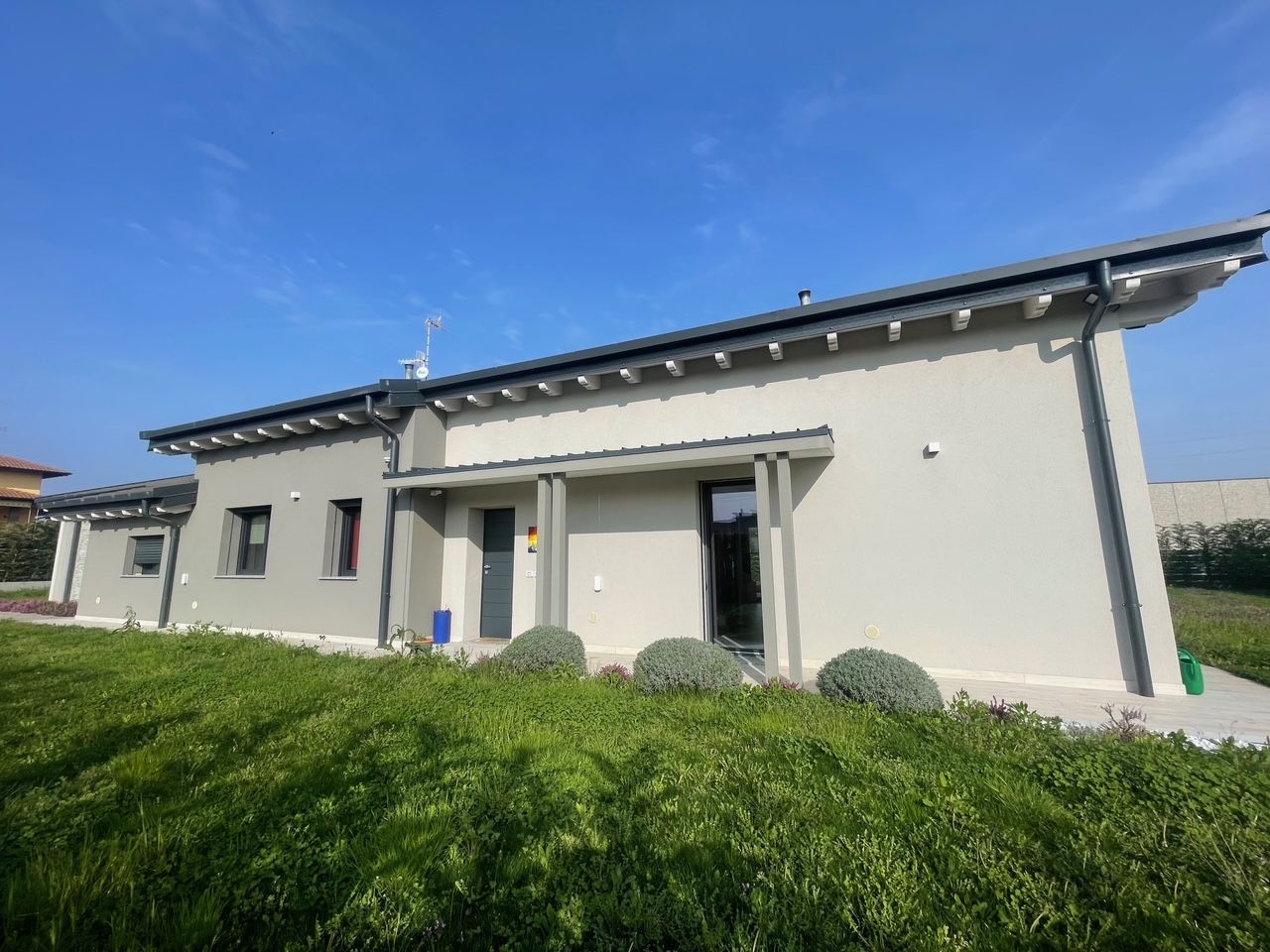 Casa moderna a un solo piano, bianca, con tetto grigio e prato verde sotto un cielo azzurro.