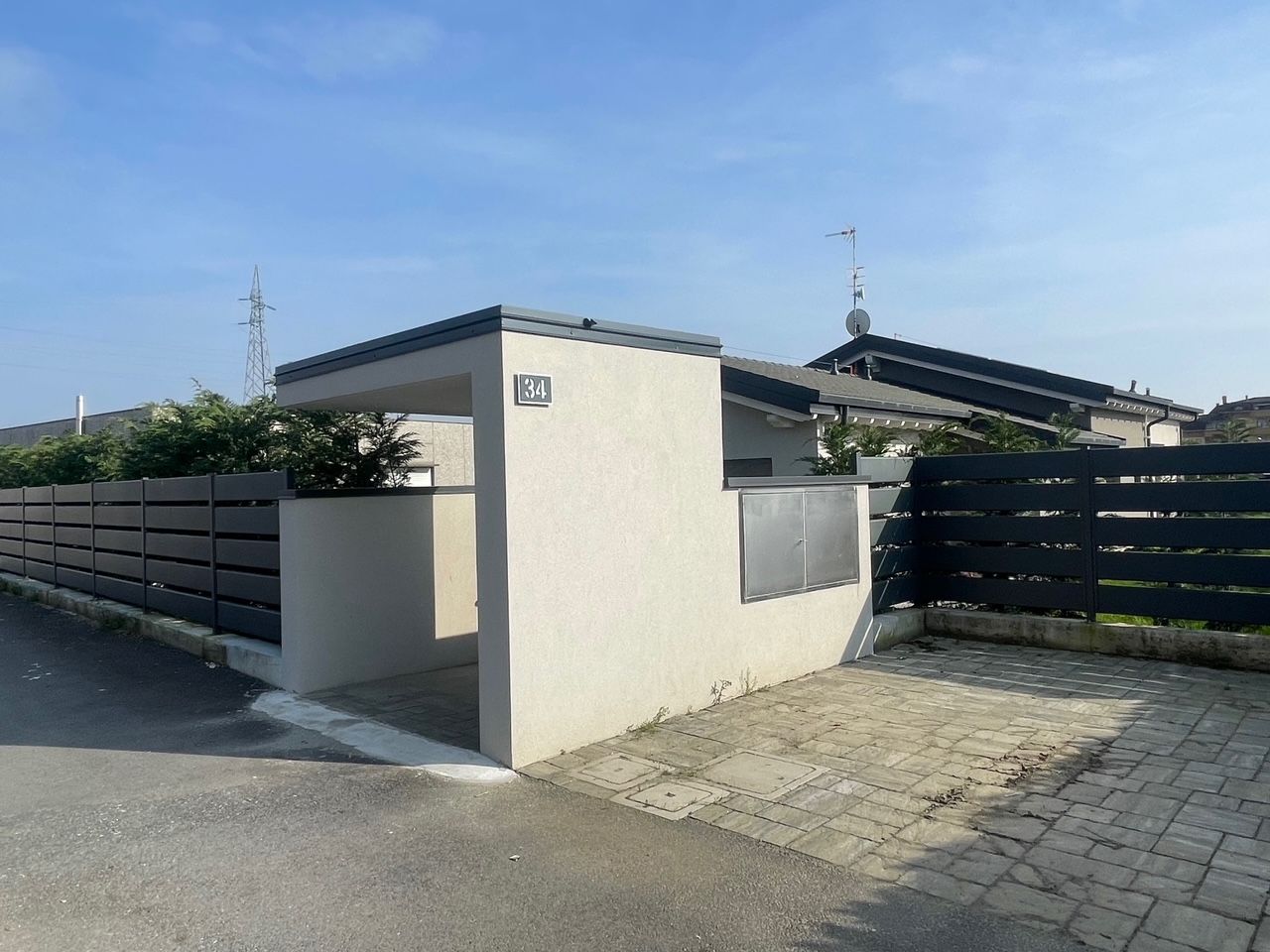 Ingresso di casa moderna con pareti color crema, recinzione scura e vialetto pavimentato sotto un cielo luminoso