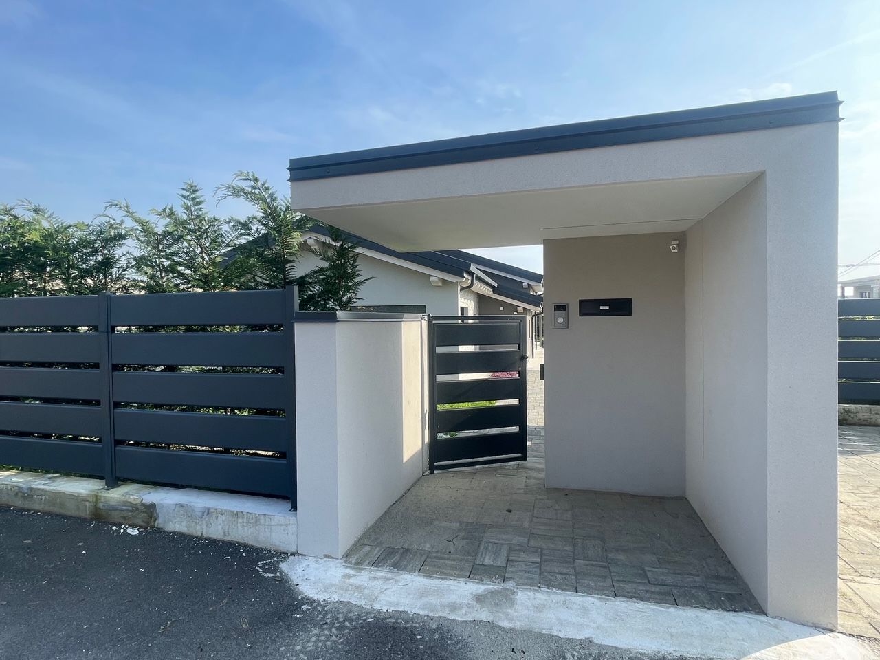 Ingresso di una casa moderna con pareti bianche, cancello scuro e vialetto piastrellato sotto un cielo limpido