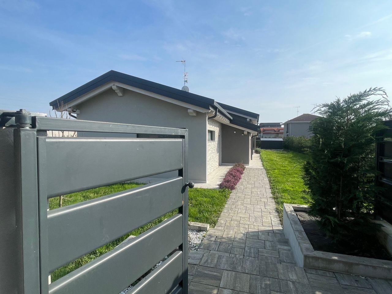 Vialetto di accesso a una residenza privata con cancello grigio, sentiero in pietra e una casetta sotto un cielo azzurro.