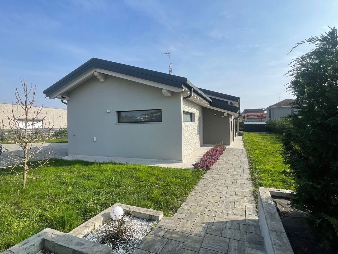 Casa a un solo piano con muri grigi, un vialetto in pietra e un prato verde sotto un cielo azzurro.