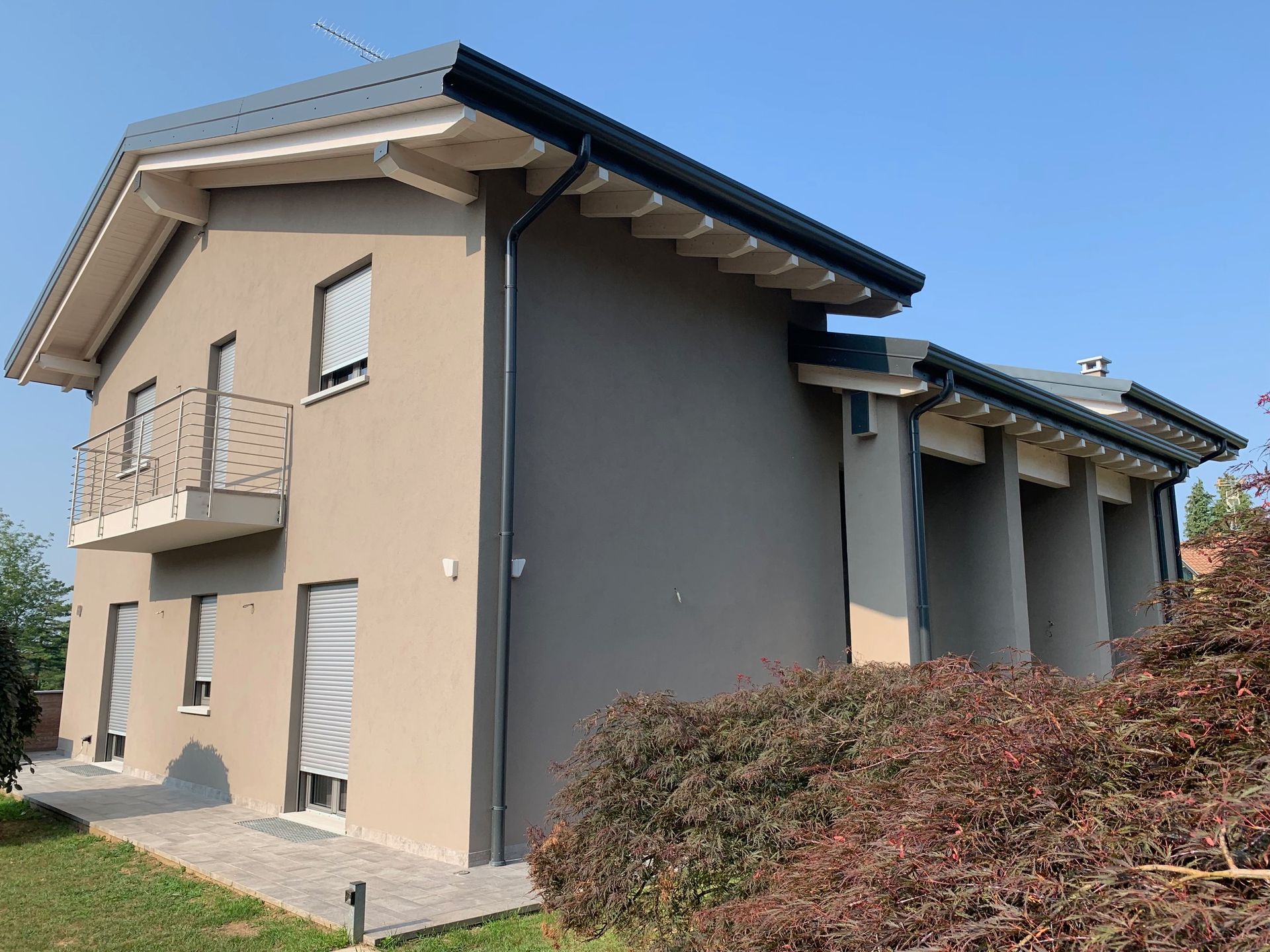 Casa beige a due piani con dettagli grigi, balcone e arbusti sotto un cielo azzurro limpido.