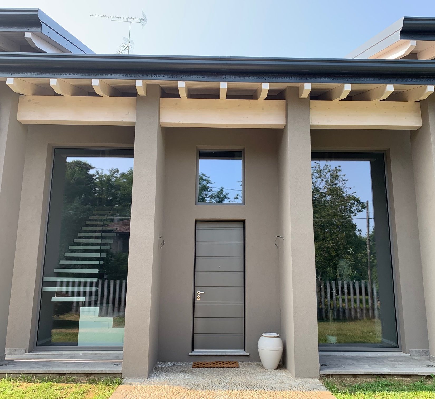 Facciata di una casa moderna con pareti beige, ampie finestre, una porta centrale e un piccolo ingresso principale.