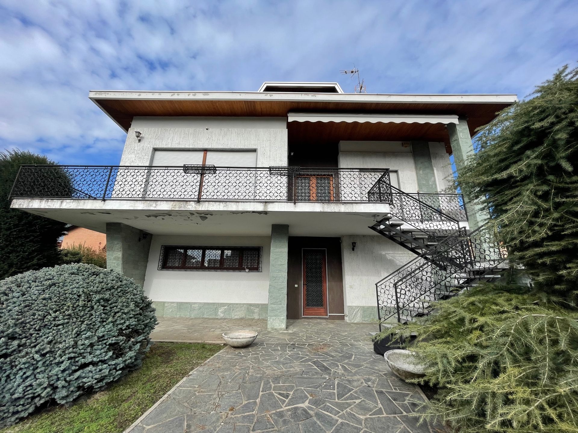 Casa a due piani con balcone, vialetto in pietra e arbusti sotto un cielo azzurro.