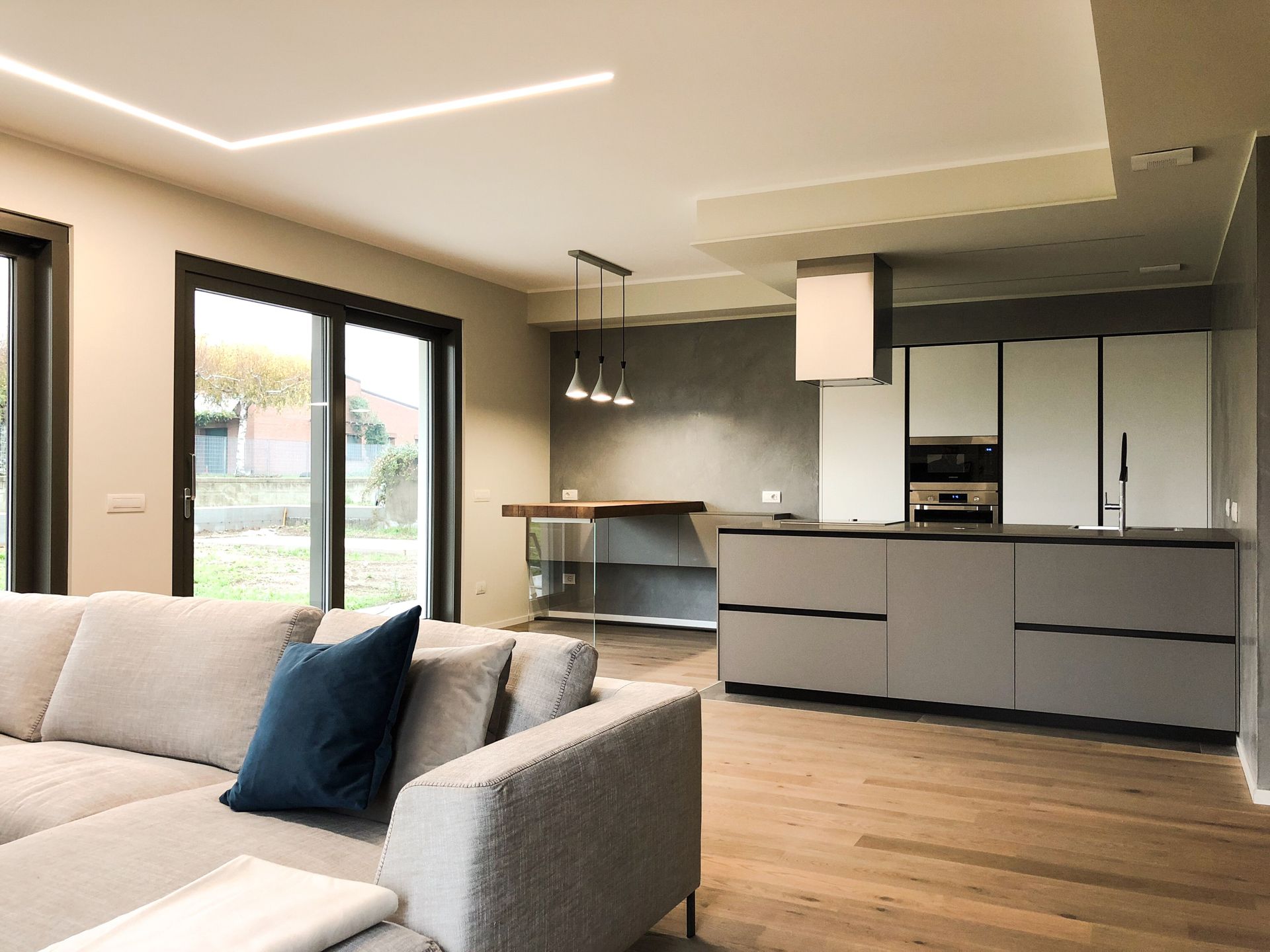 Soggiorno e cucina moderni a pianta aperta con divano grigio, pavimento in legno, porte con cornice nera e lampade a sospensione.