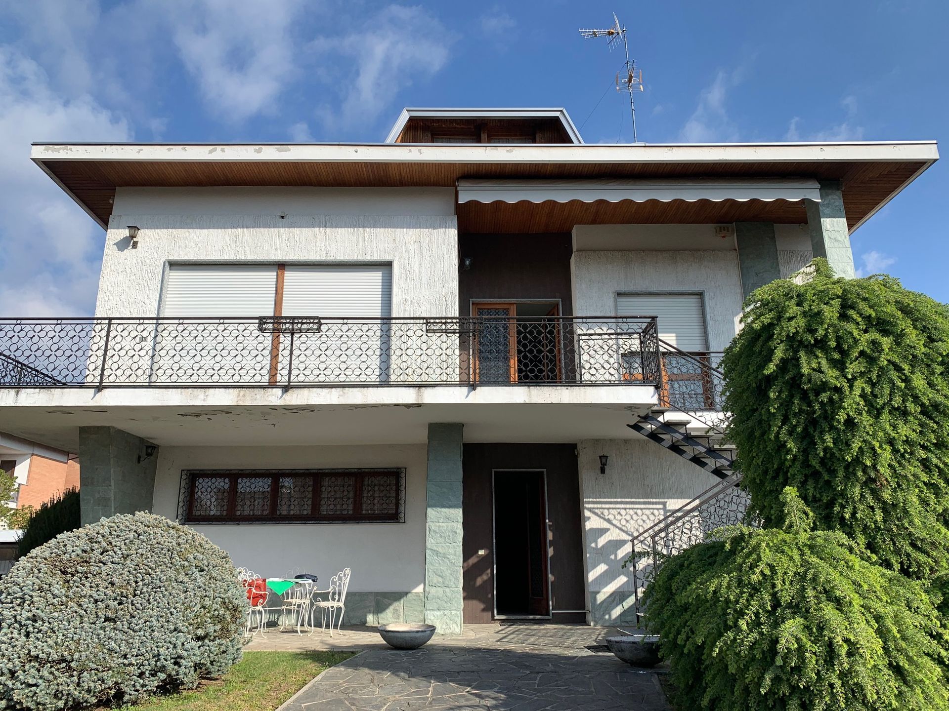 Casa moderna a due piani con balcone, facciata bianca e un piccolo giardino sotto un cielo azzurro.