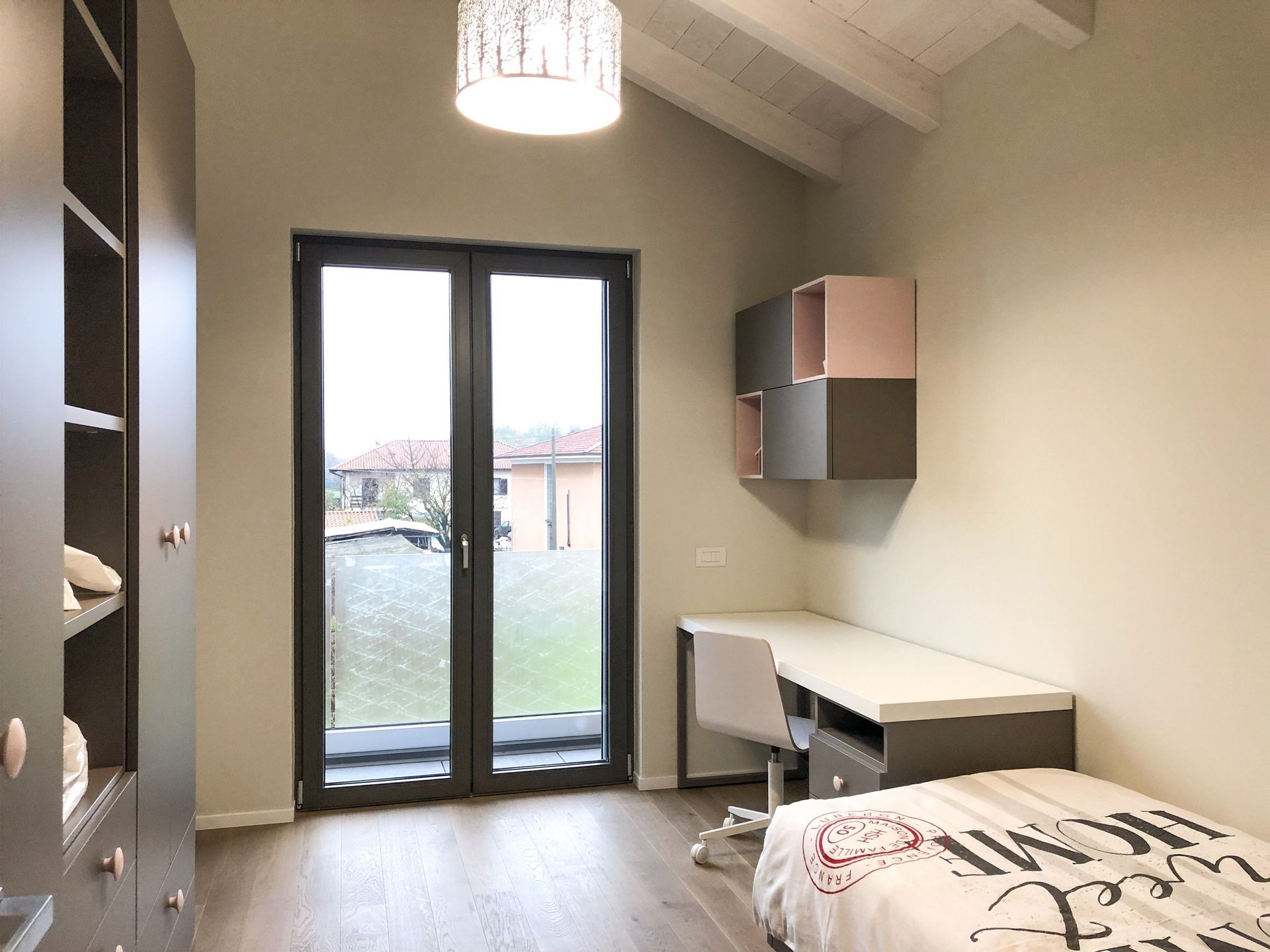 Piccola camera da letto con scrivania, armadi a muro, letto e alte porte finestre che si aprono verso l'esterno.