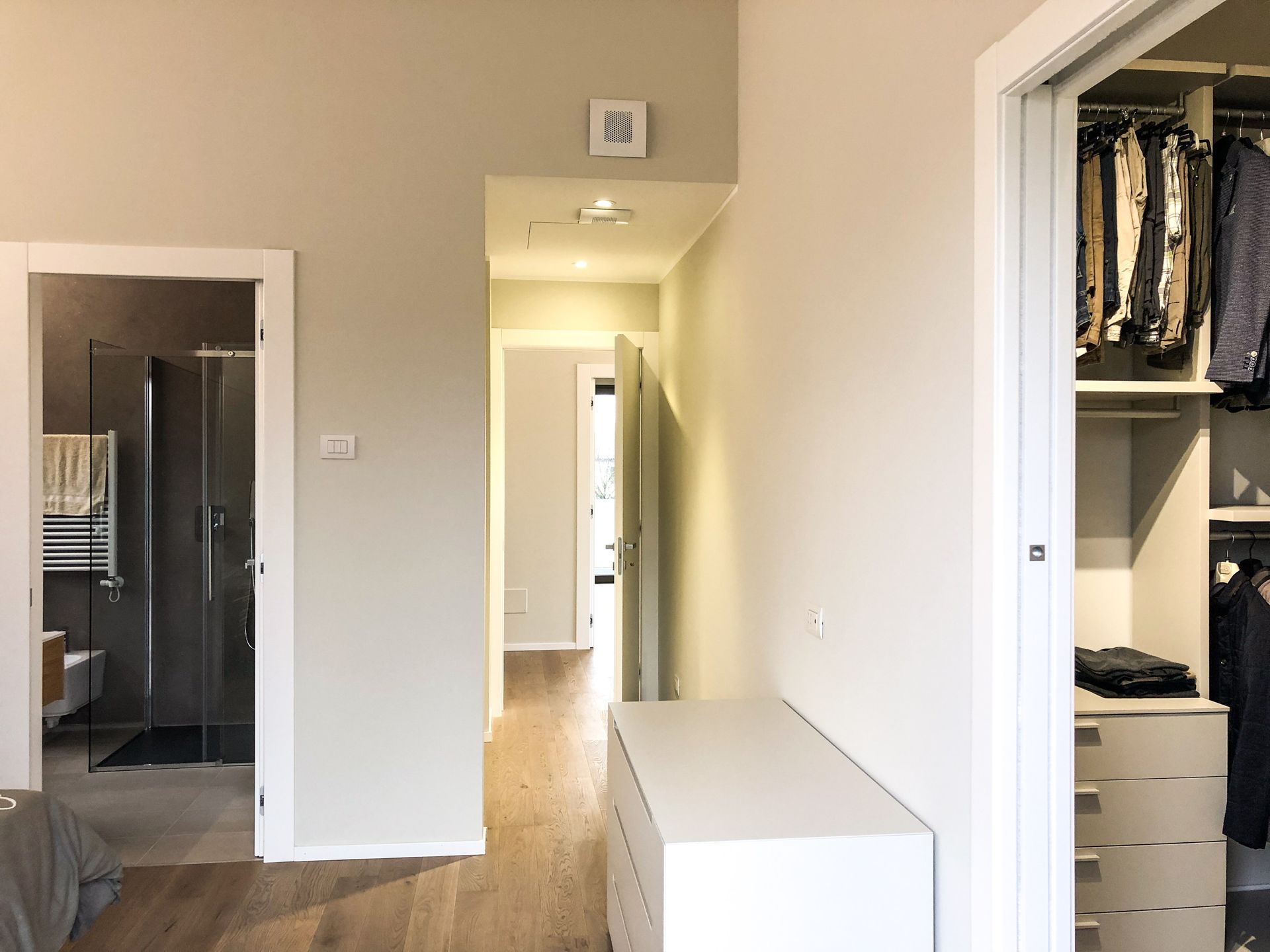 Corridoio moderno con finiture bianche, pavimento in legno, bagno e cabina armadio a vista e un mobiletto bianco.