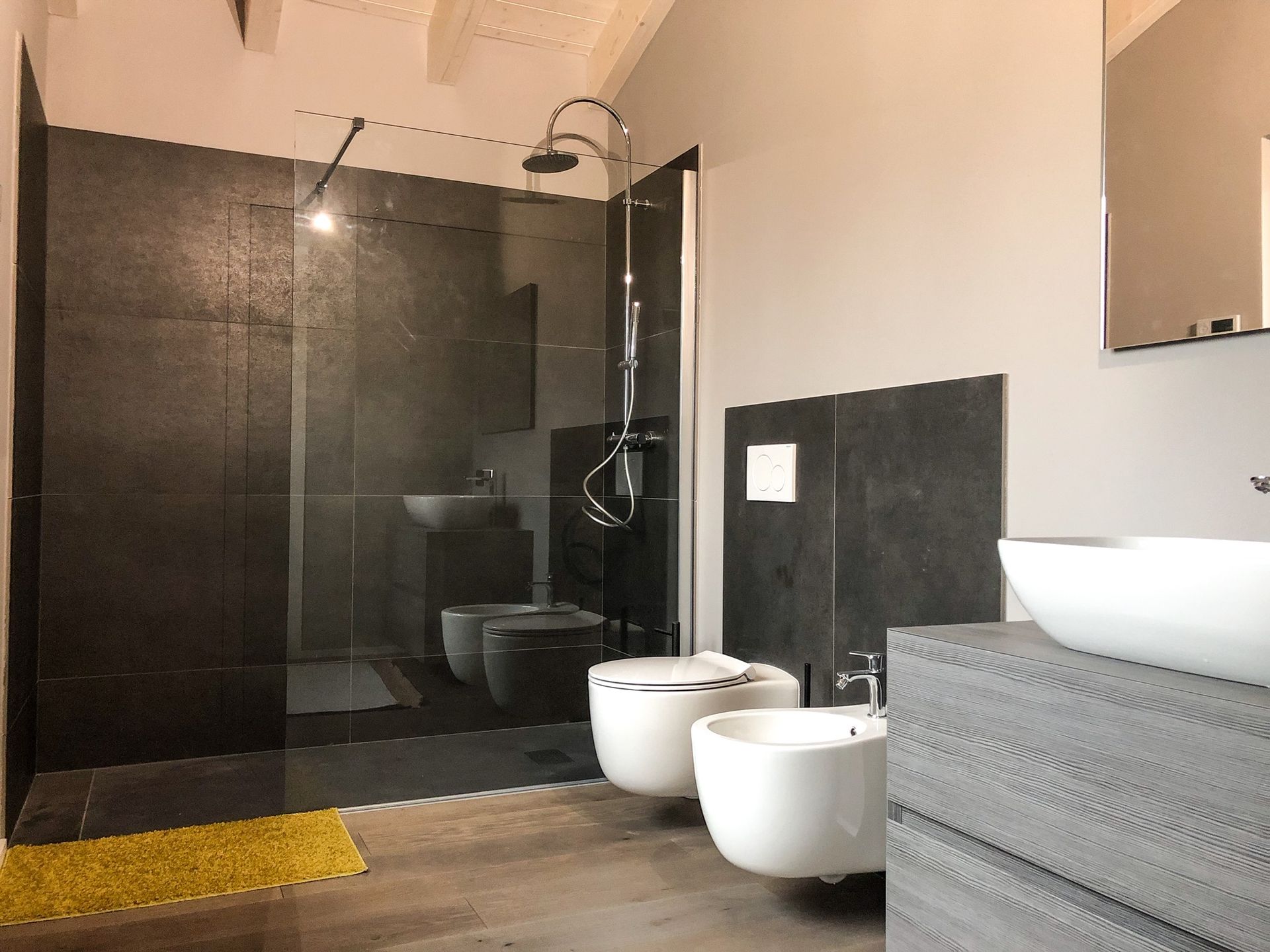 Bagno moderno con doccia piastrellata di nero, box doccia in vetro e due lavabi a ciotola bianchi su un mobiletto grigio.