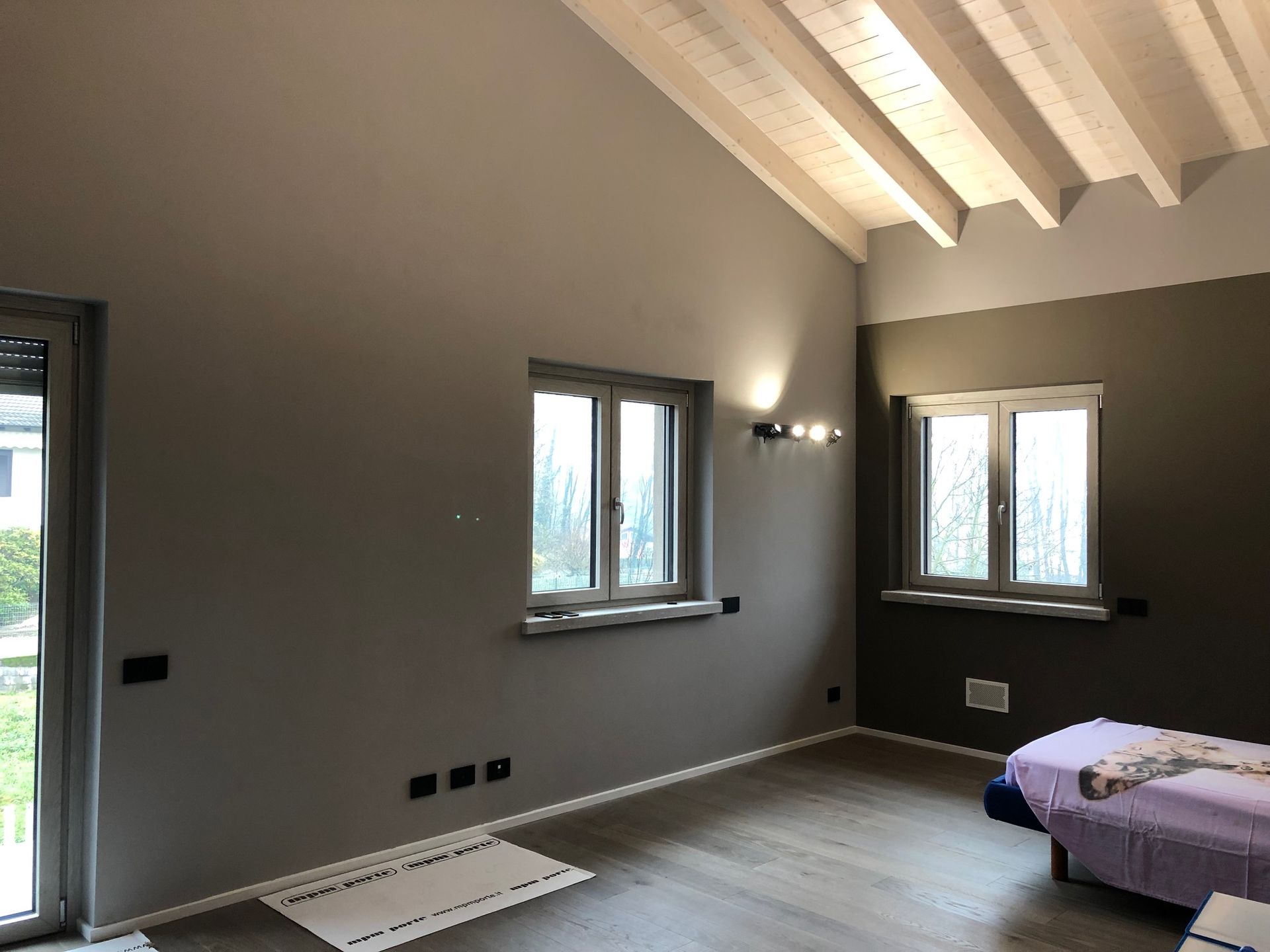 Camera da letto vuota con pareti grigie, travi a vista, tre finestre e un letto in un angolo.