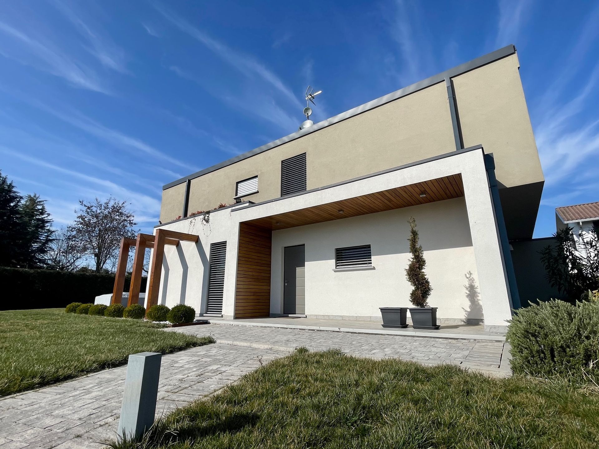 Casa moderna a due piani con patio bianco, pergola in legno e prato in pendenza sotto un cielo azzurro.