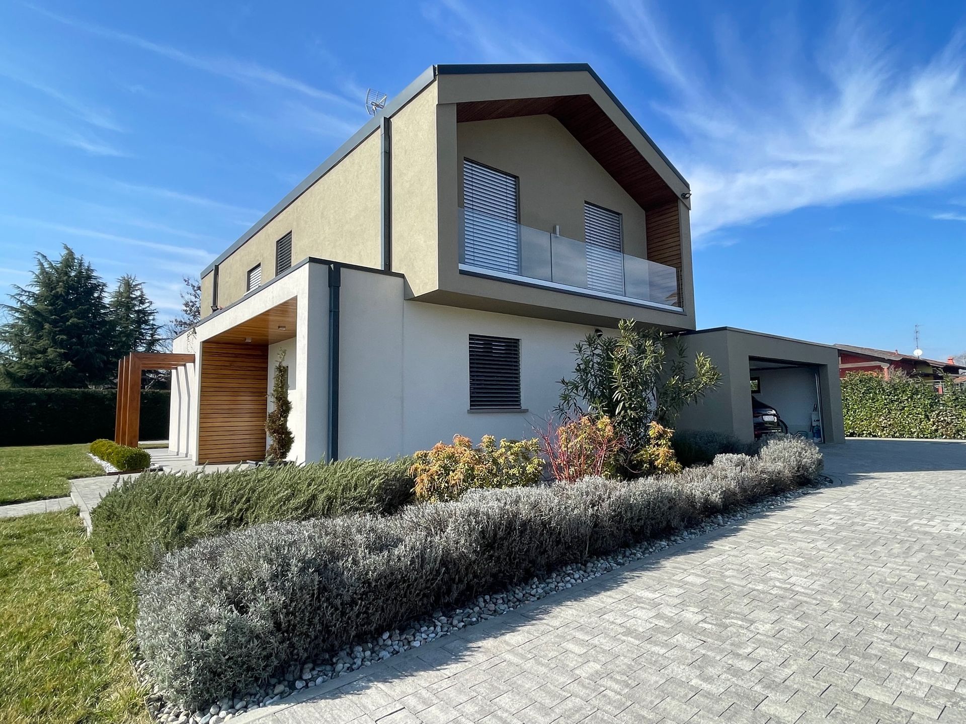 Casa moderna a due piani con balcone, pareti in stucco bianco, garage in legno e vialetto pavimentato sotto un cielo azzurro.