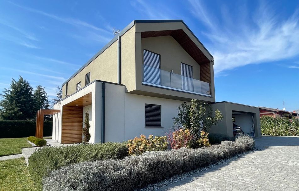 Casa moderna a due piani con balcone, pareti bianche, dettagli in legno e un giardino anteriore curato sotto un cielo azzurro.