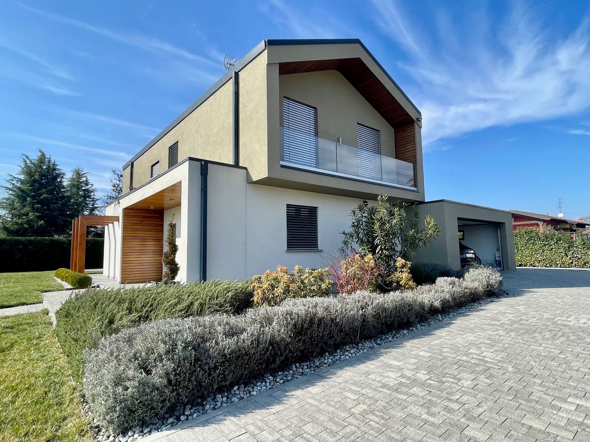 Casa moderna a due piani con balcone, vialetto d'accesso e giardino paesaggistico sotto un cielo azzurro.