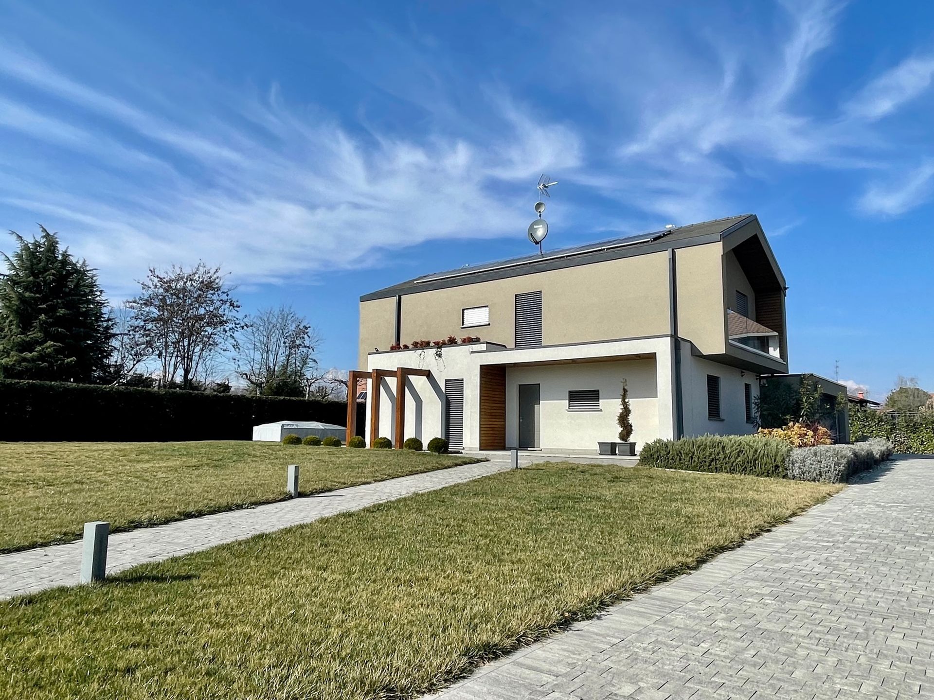 Casa moderna a due piani con prato in pendenza, vialetto in cemento e cielo azzurro in una giornata di sole.