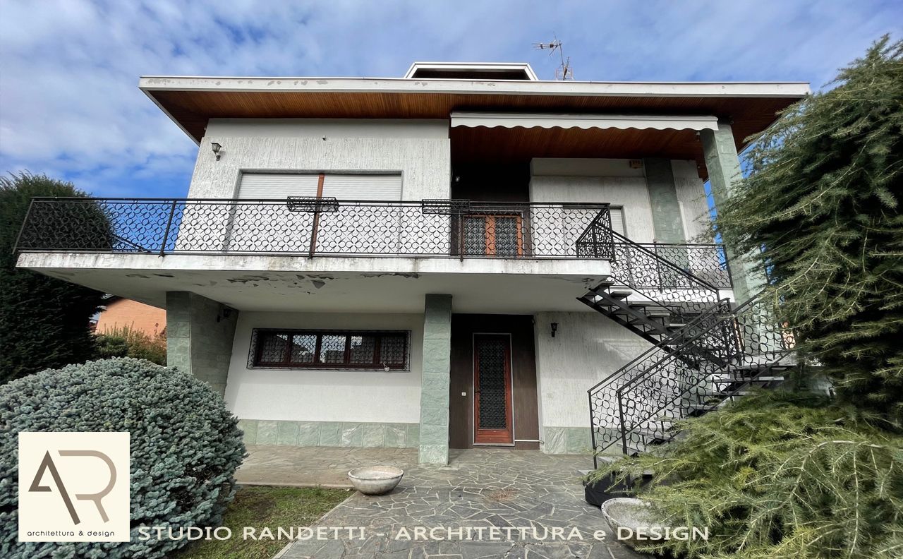 Casa bianca a due piani con balcone, scala esterna e dettagli rosso-marroni, situata dietro un cancello.