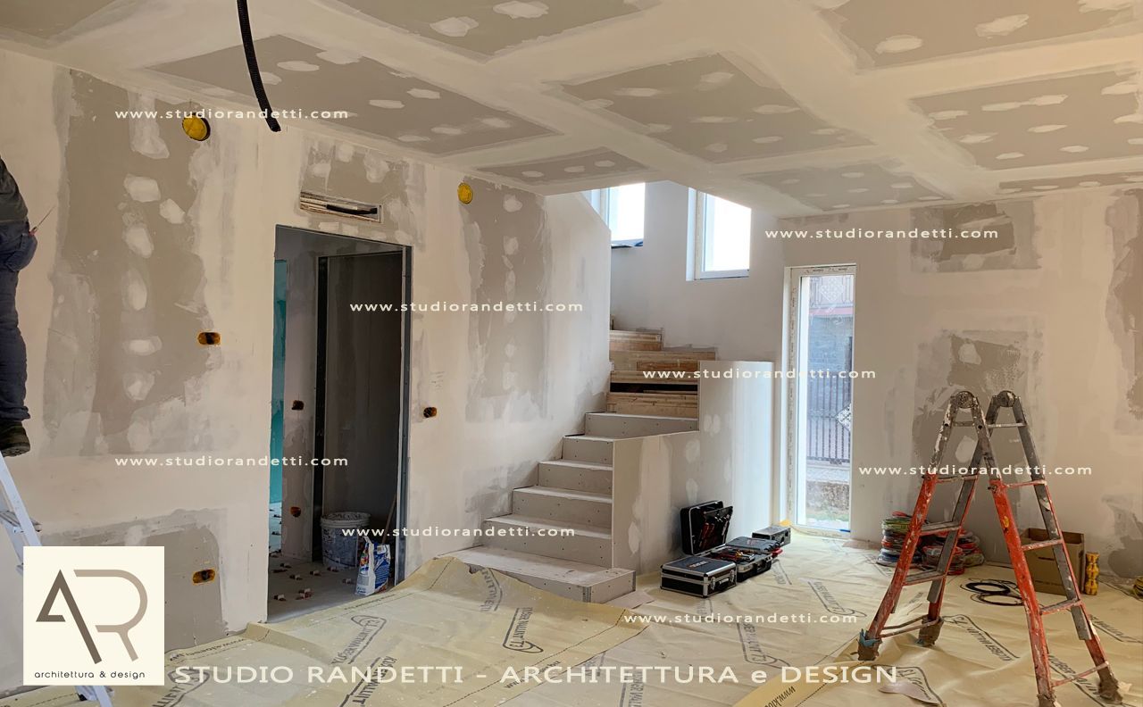 Cantiere in fase di ristrutturazione con pareti in cartongesso bianco, scala, scaletta e porta ad arco non rifinita in una stanza luminosa.