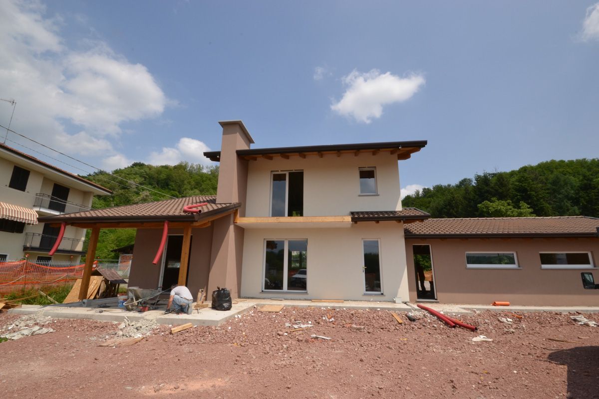 Casa beige a due piani con portico piastrellato di rosso e cortile di terra battuta non rifinito sotto un cielo azzurro.