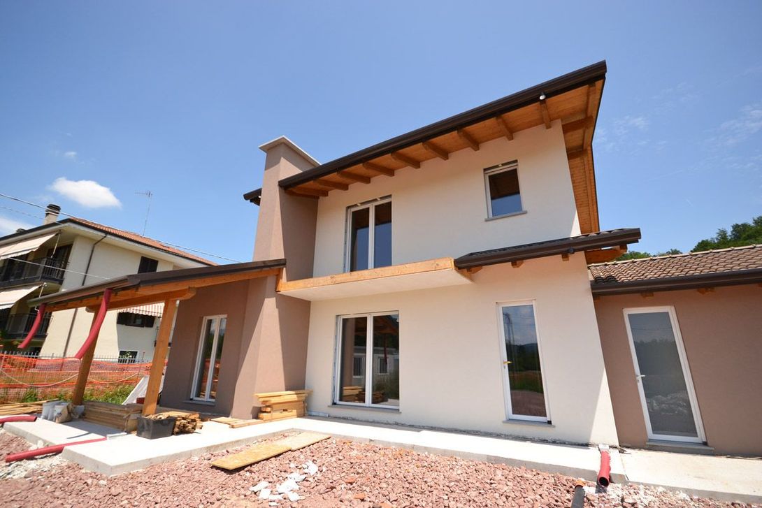 Esterno di una moderna casa a due piani con stucco beige, ampie finestre e tetto di tegole sotto un cielo azzurro.