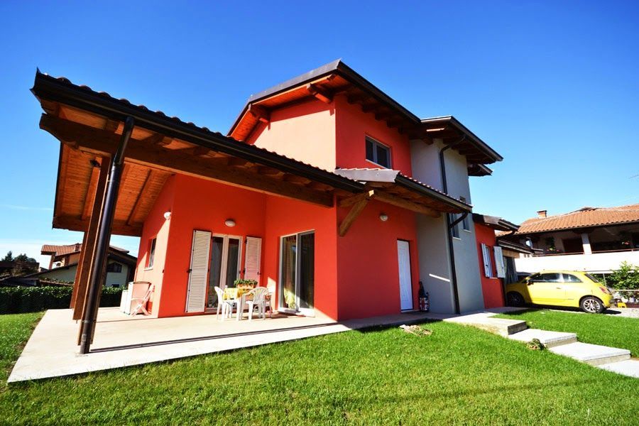 Casa moderna a due piani di colore rosso con portico coperto, prato verde e cielo azzurro brillante.