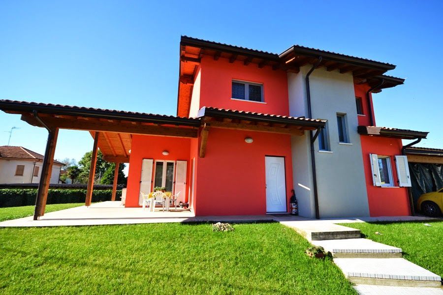 Casa moderna rosso-arancio con patio coperto e prato verde sotto un cielo azzurro brillante
